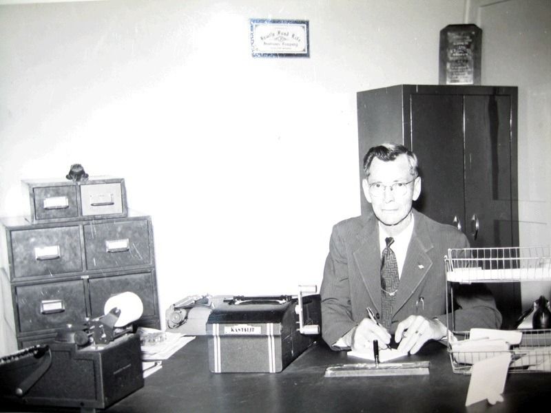 Man seated at a desk in an office, with files, typewriter, and cabinets behind him.