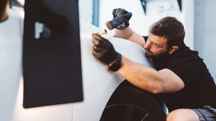 Paintless Dent Repair Person using a hammer to repair a white car panel, focused close-up in a garage.