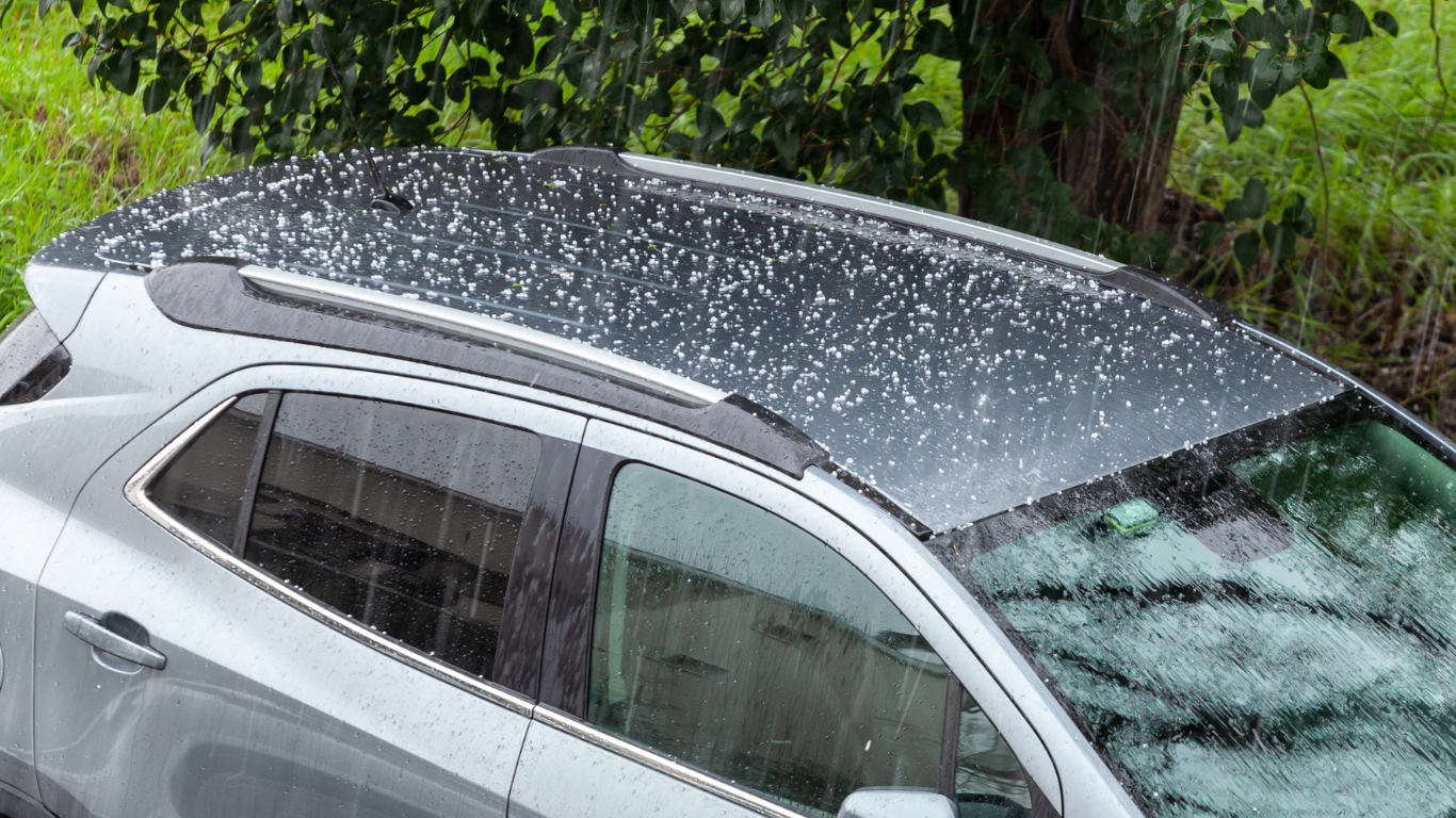 Auto Hail Damage Repair Silver car roof covered in white hail or snow, parked beside green bushes