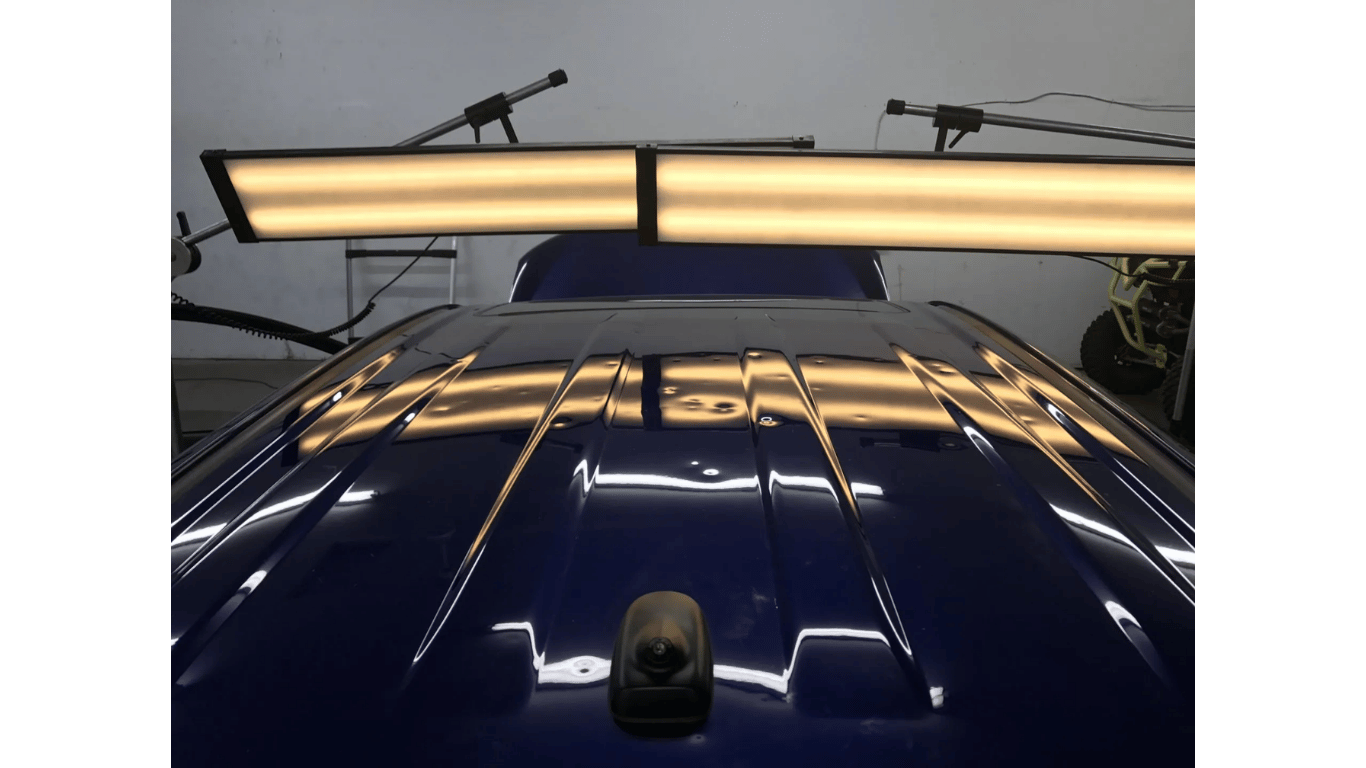 Blue car hood under bright overhead lights in a garage, with reflections and a roof-mounted camera.