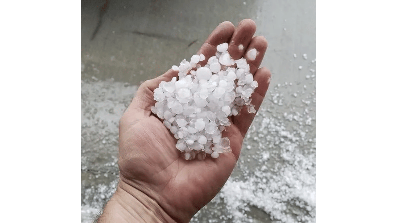 A hand holding a handful of small white hailstones, with more hail scattered on the ground.