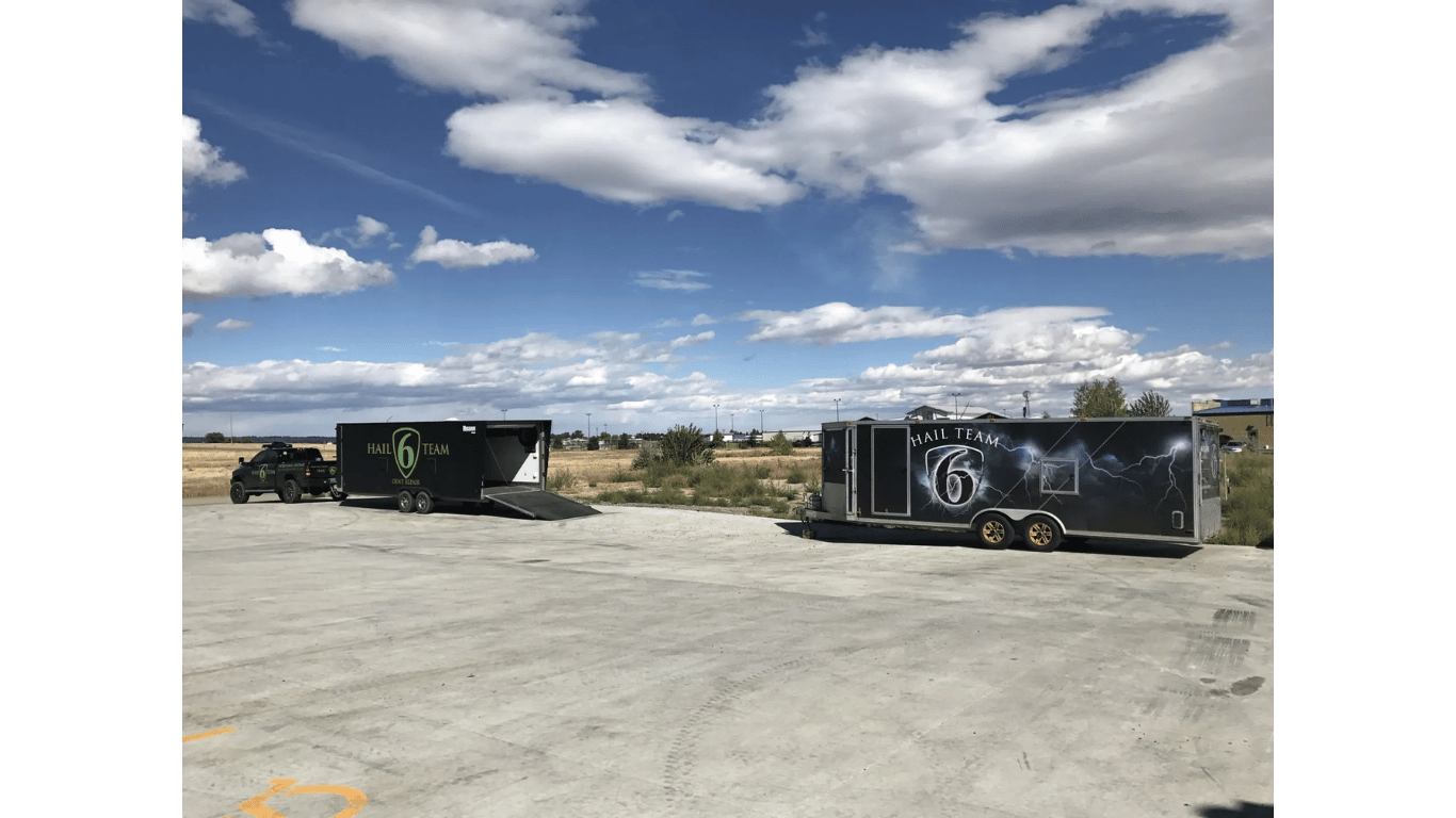 Two black dump trailers parked on a wide gravel lot under a bright blue sky with clouds