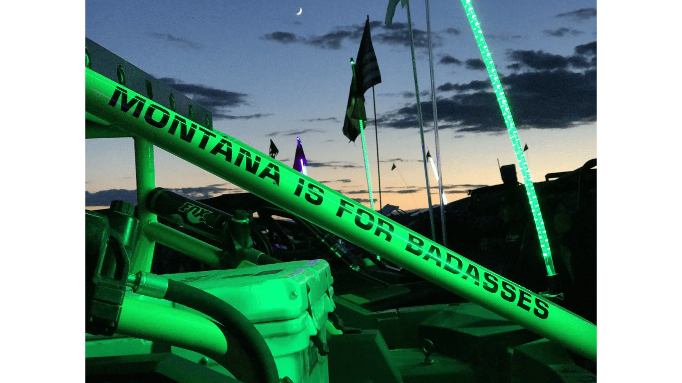 Green-lit truck with “Montana is for badasses” banner at dusk, with flags and poles in the background