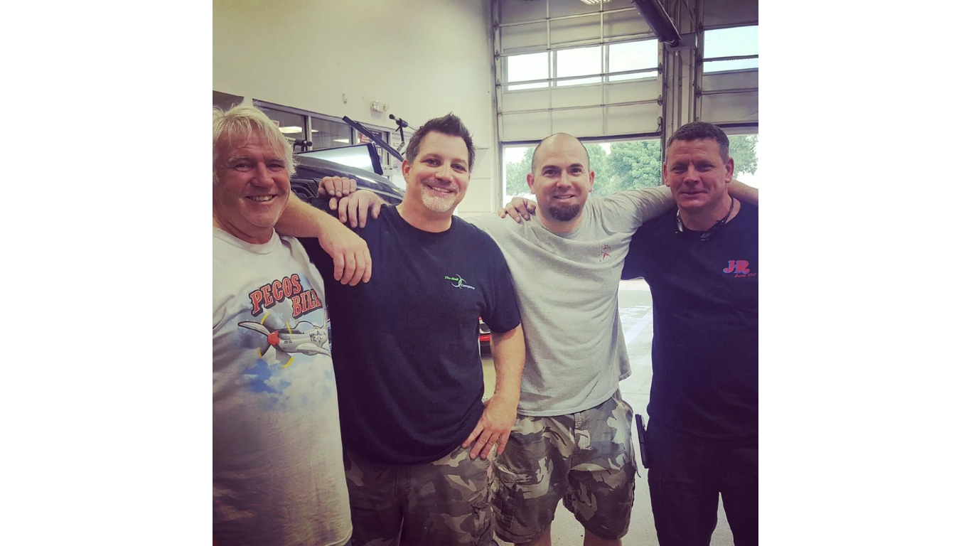 Four smiling people standing together indoors, arms around each other in a warehouse-like space.