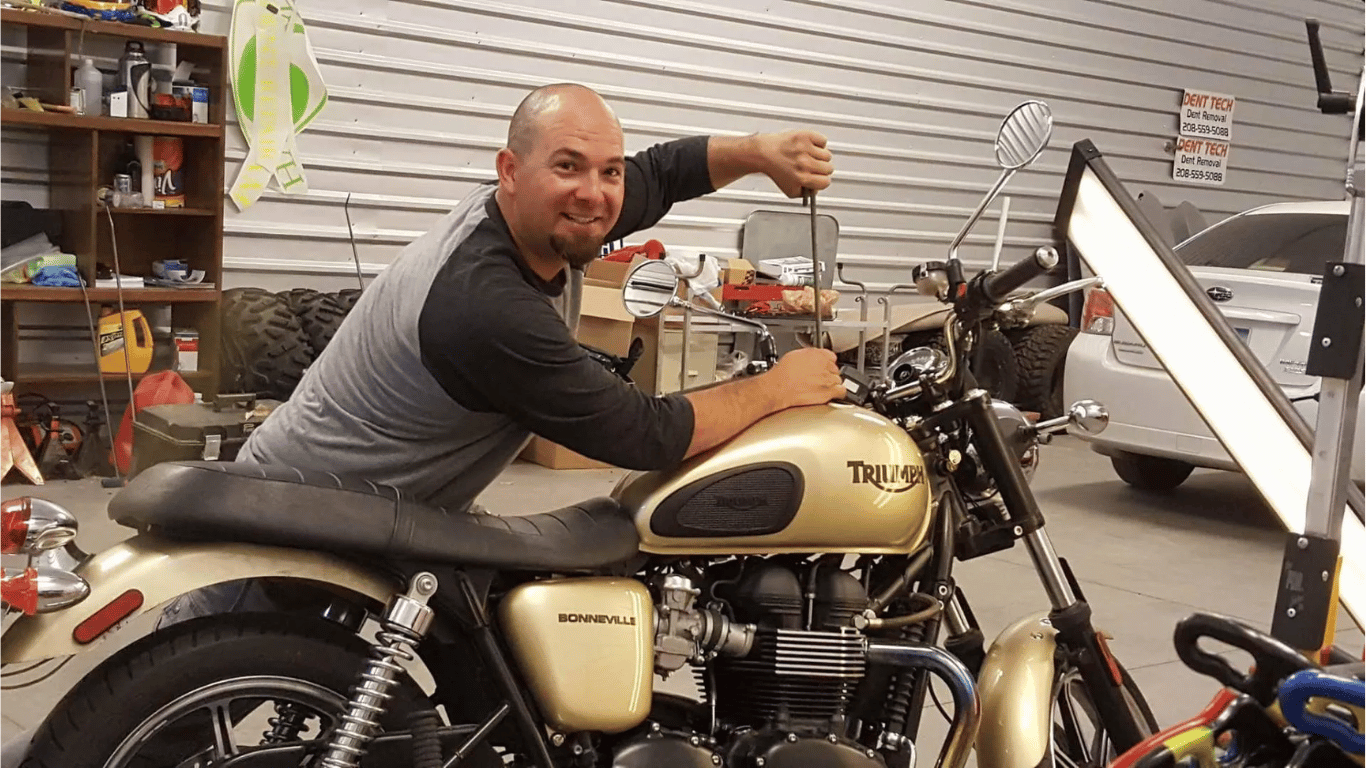 Man smiling on a beige motorcycle in a garage, pointing toward the handlebars