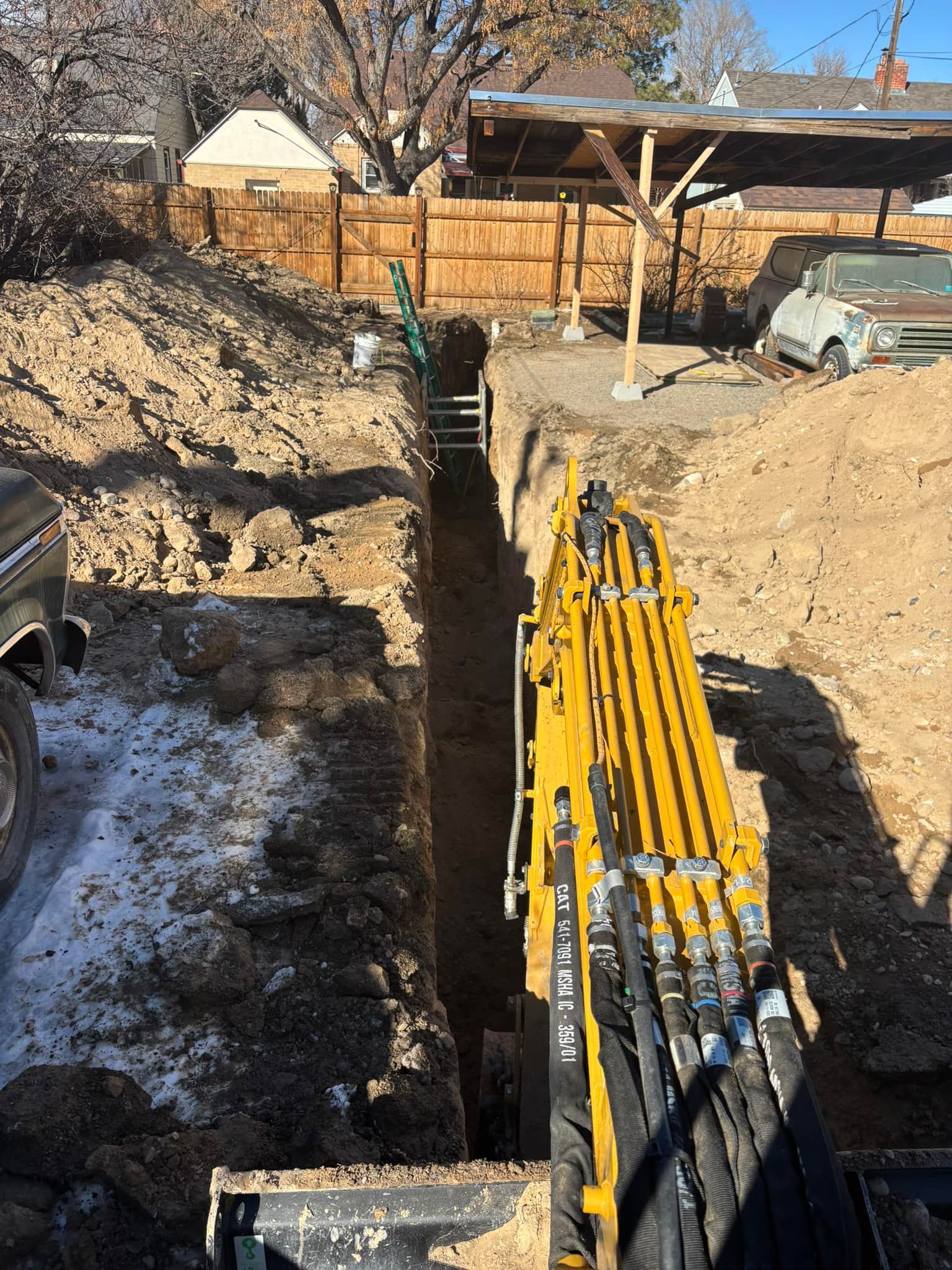 Yellow excavator digging a long, narrow trench in a backyard with a wooden fence and structure in the background.