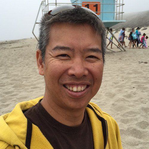 A man wearing sunglasses and a yellow jacket is smiling on the beach