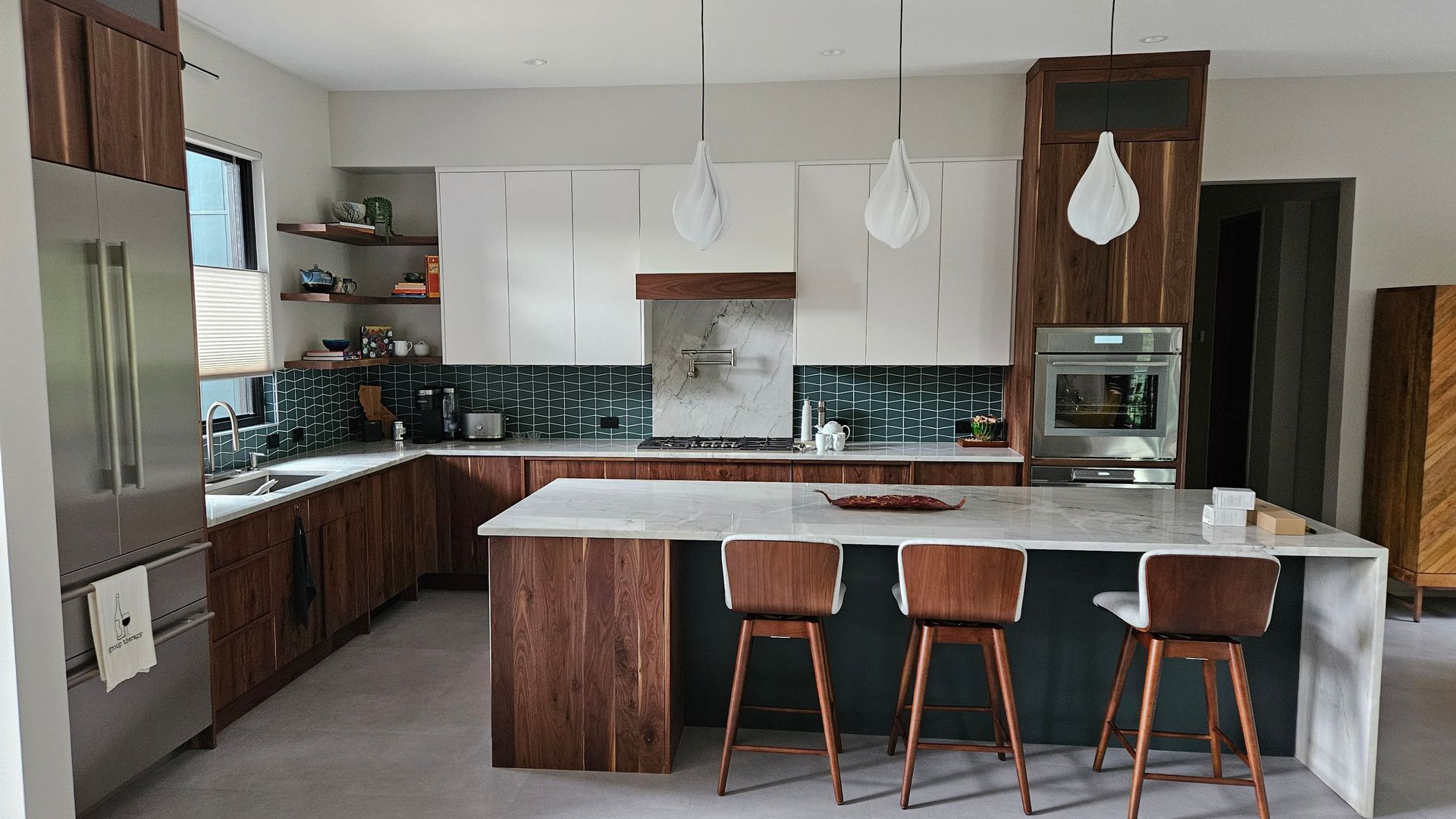 Modern kitchen with wood cabinets, white island, and three stools, lit by pendant lights.