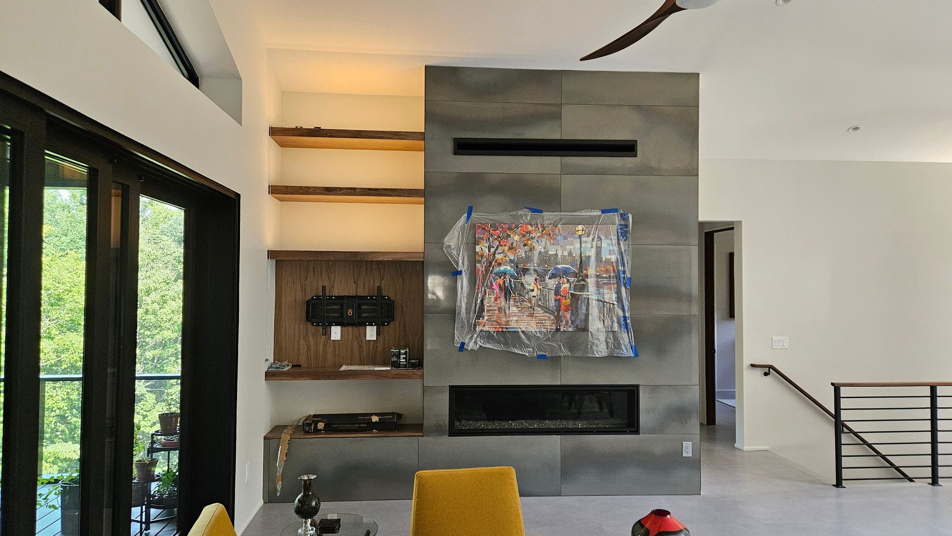 Modern living room with a gray fireplace, abstract art, yellow chair, and large windows overlooking greenery