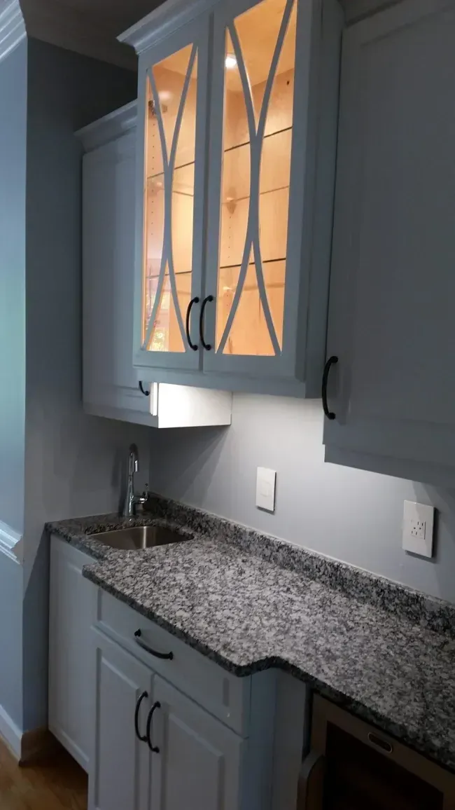 Gray kitchen cabinets and granite countertop with a lit glass-front upper cabinet and sink.