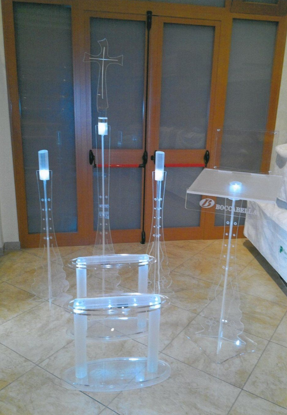 Arredi in acrilico trasparente: pulpito, sedie, podi con accenti luminosi, inseriti in una stanza con porte in legno.