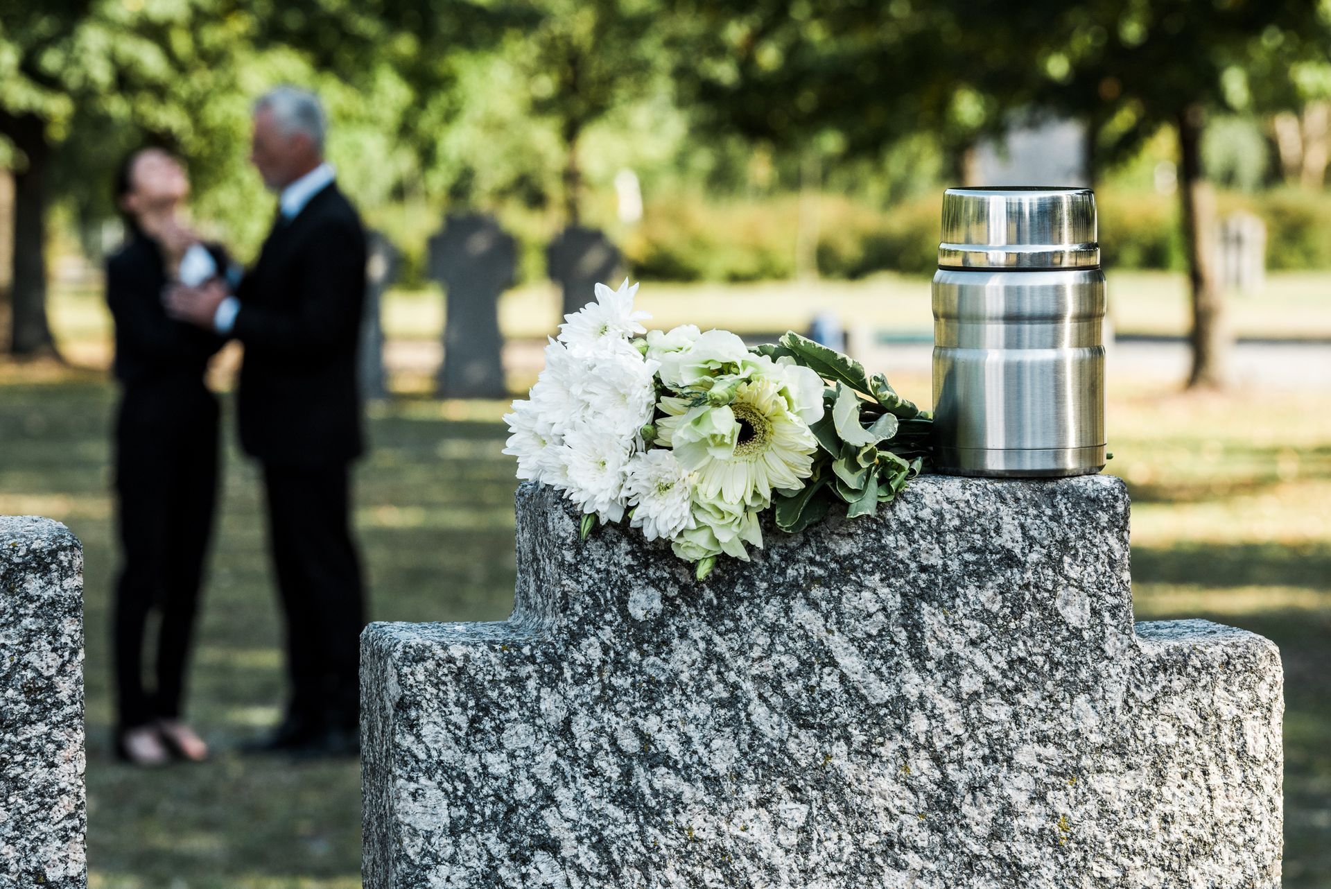 Lapide con fiori e un'urna in un cimitero, sullo sfondo persone in lutto.