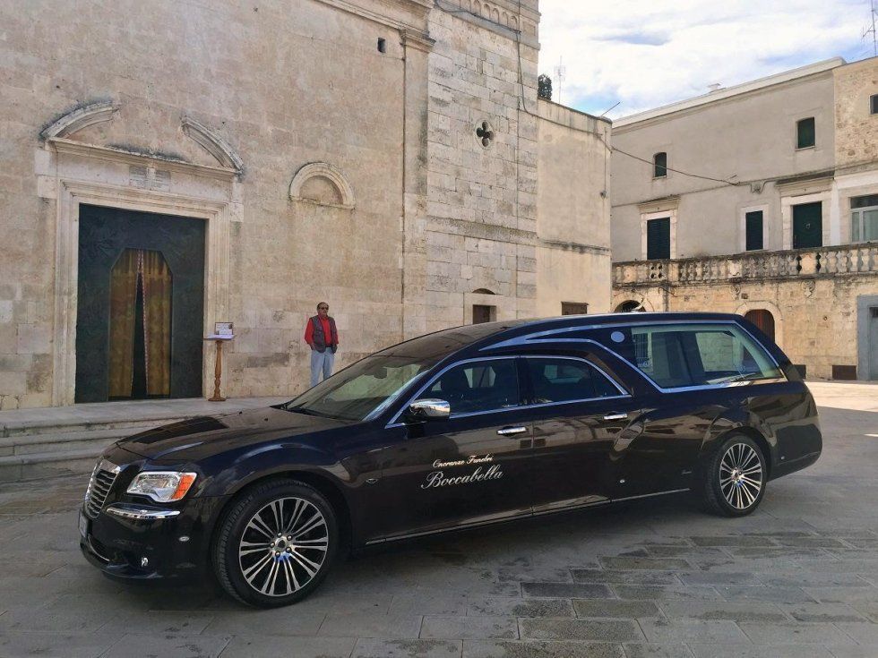 Un carro funebre nero è parcheggiato davanti a una chiesa in pietra; una persona è in piedi vicino all'ingresso.