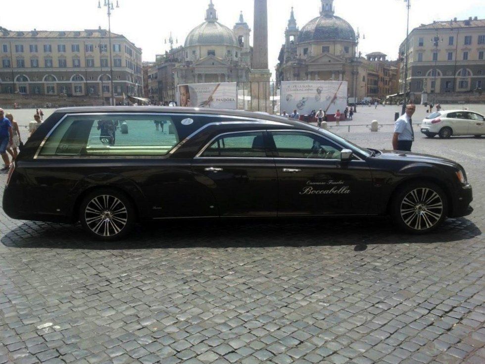 Un carro funebre scuro parcheggiato in una piazza acciottolata, con la Basilica di San Pietro visibile sullo sfondo.
