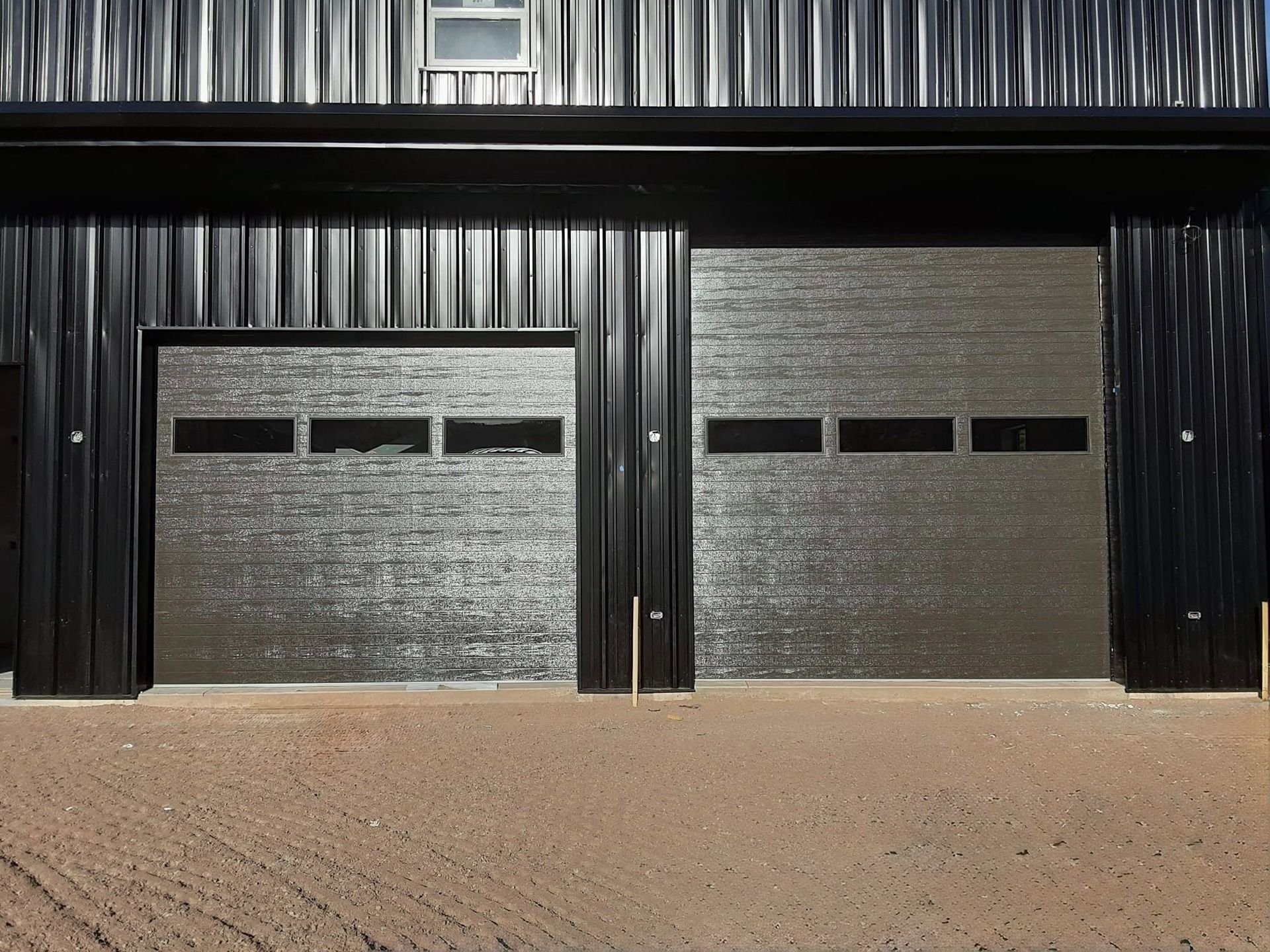 Two dark metal garage doors with rectangular windows set in a black corrugated metal building.