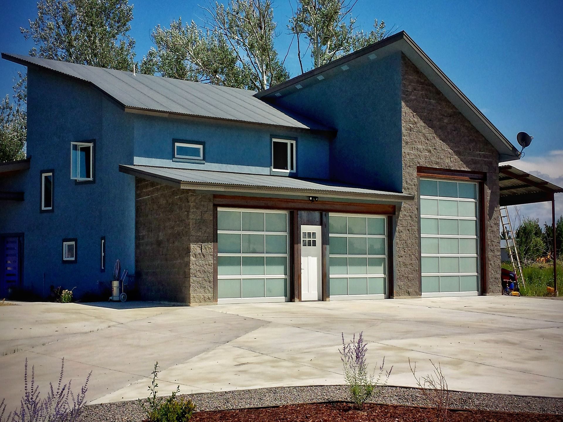 A modern house featuring blue siding, stone accents, a metal roof, and three large glass-paneled garage doors.