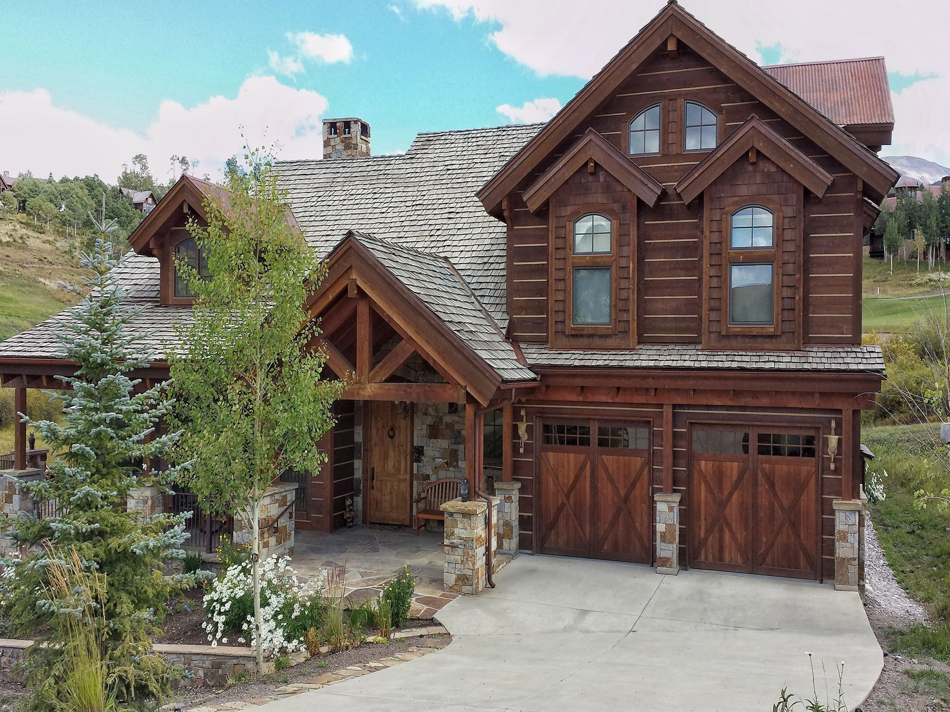 A rustic two-story wooden log cabin home with a stone foundation, gray shingled roof, and a two-car wooden garage.