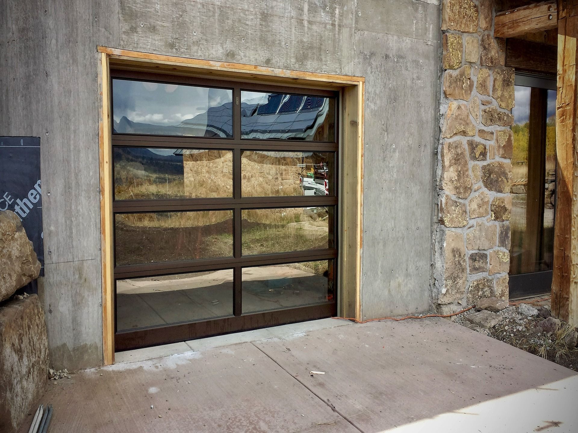 A modern glass garage door with a dark metal frame installed in a concrete wall next to a stone exterior.