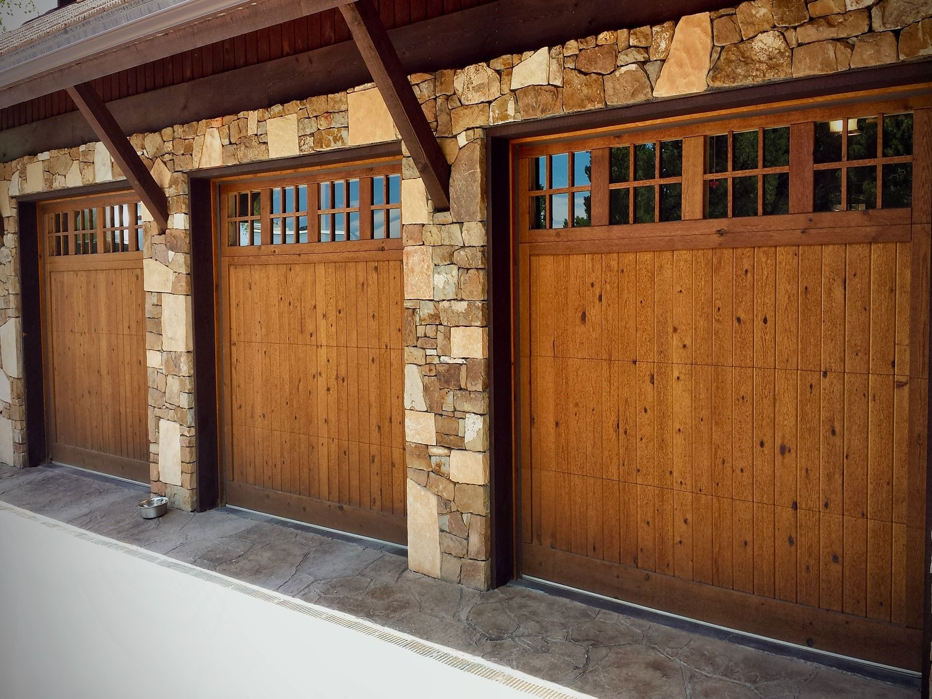 Three wooden garage doors with windows, set in a stone facade under a dark wood overhang.