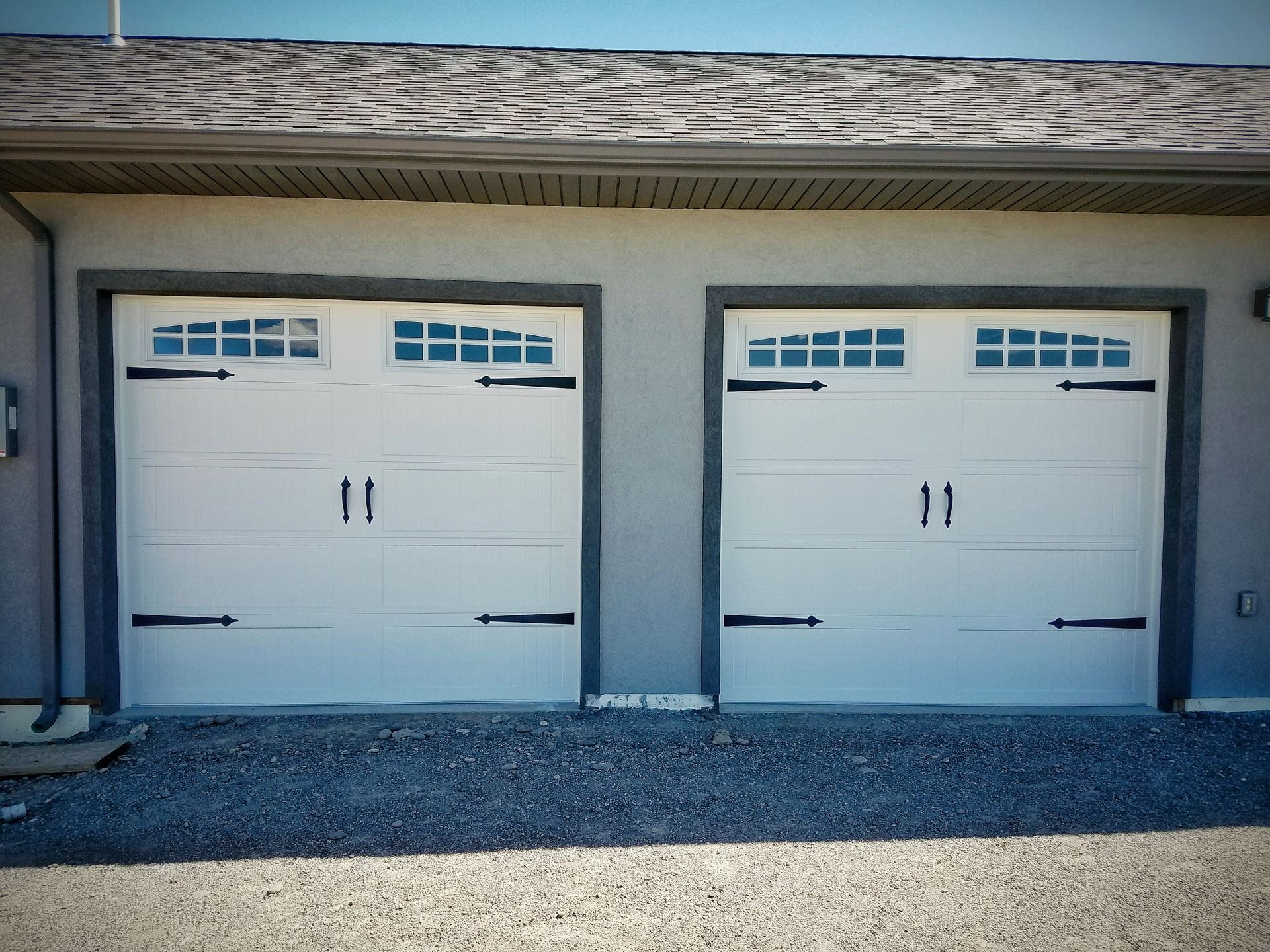 Two white garage doors with black decorative hinges, handles, and rectangular windows, set against a grey stucco wall.