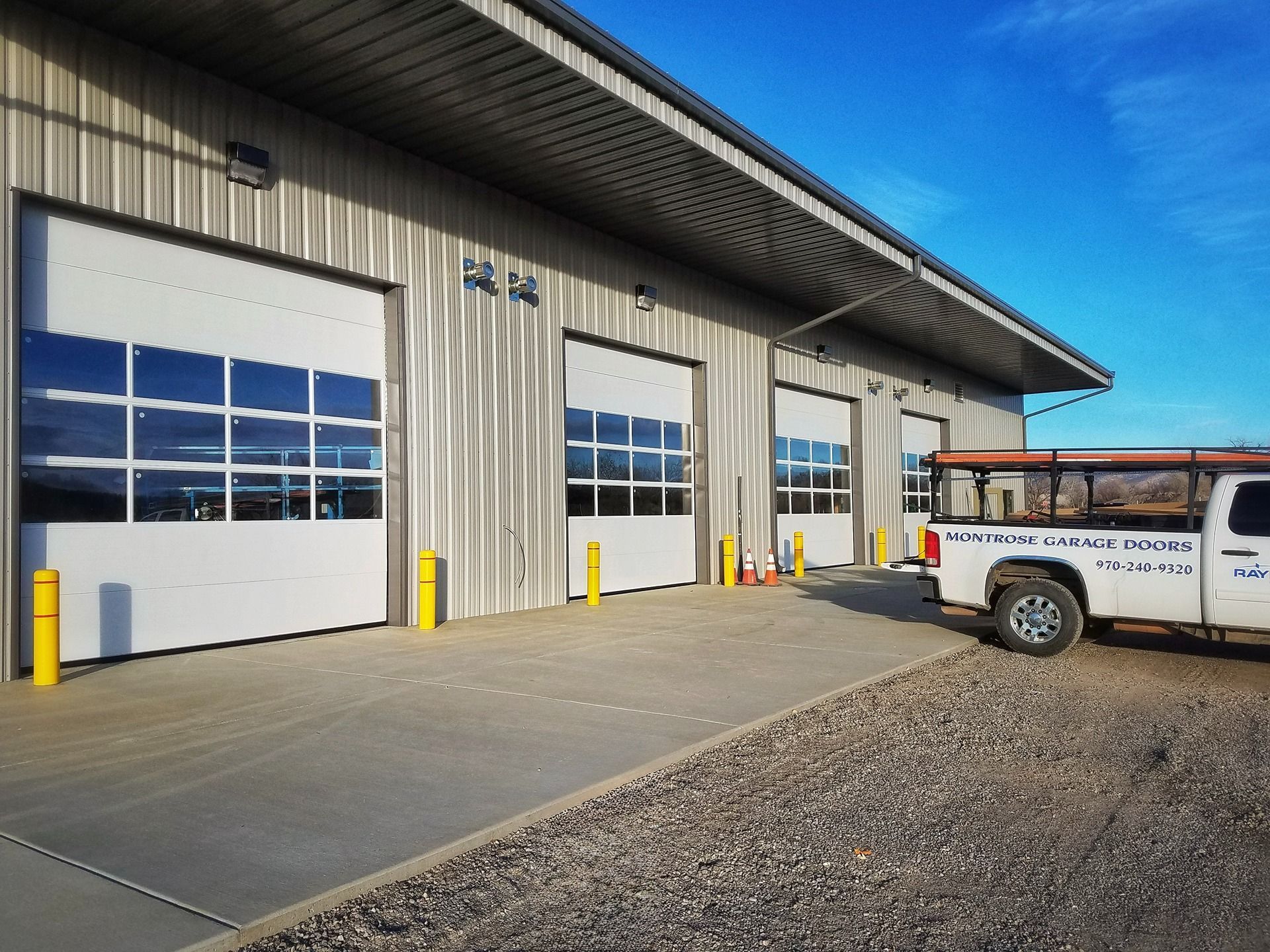 A white metal industrial building with four glass-paneled garage doors and a work truck parked on a gravel lot.