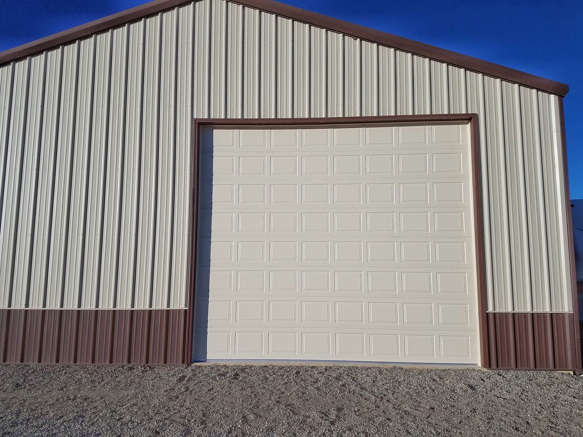 A metal workshop building with a beige garage door and tan walls with brown trim on a gravel lot under a blue sky.