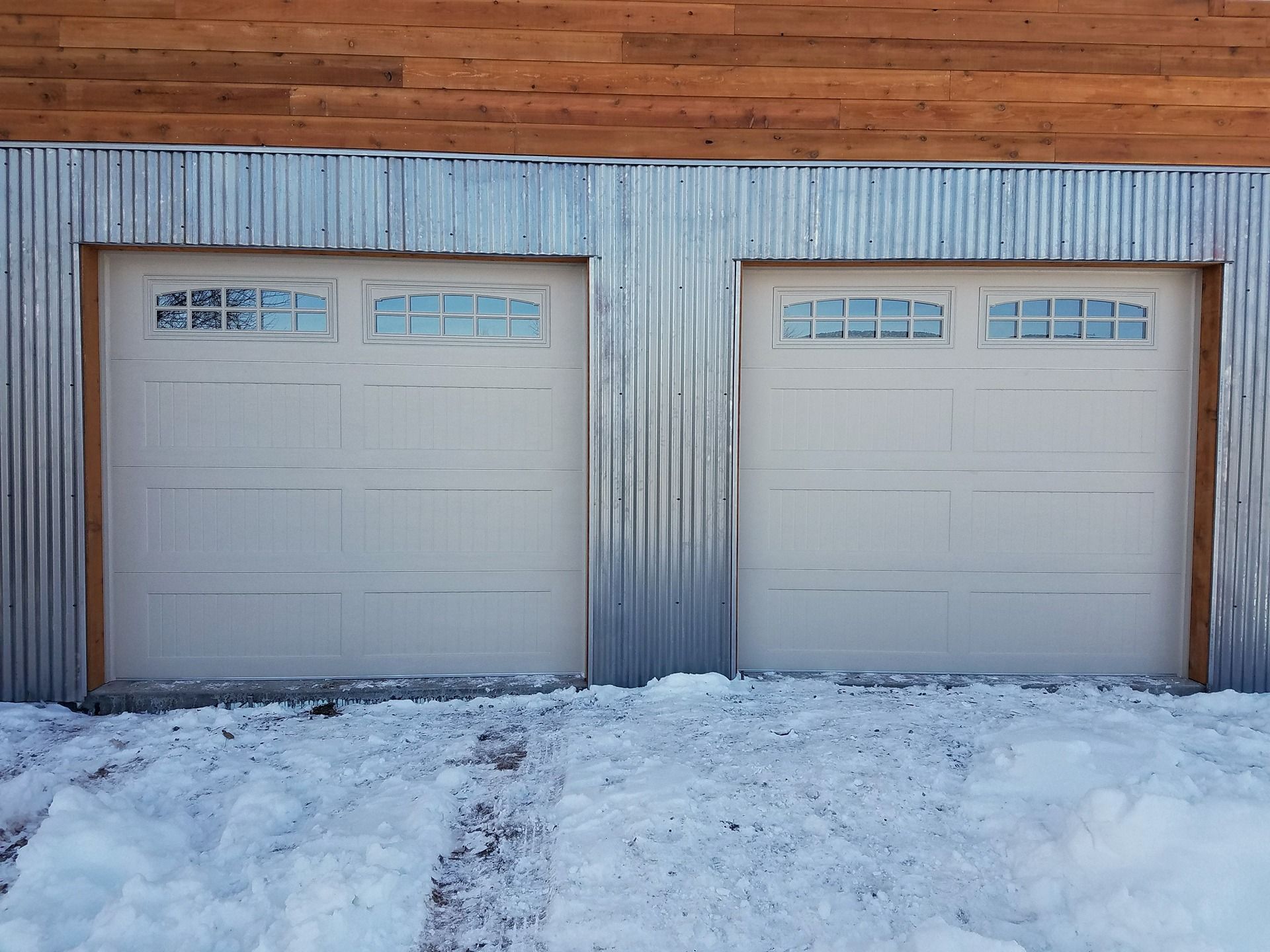 Two garage doors with window panels, framed by corrugated metal siding below a horizontal wood-paneled wall and snow.