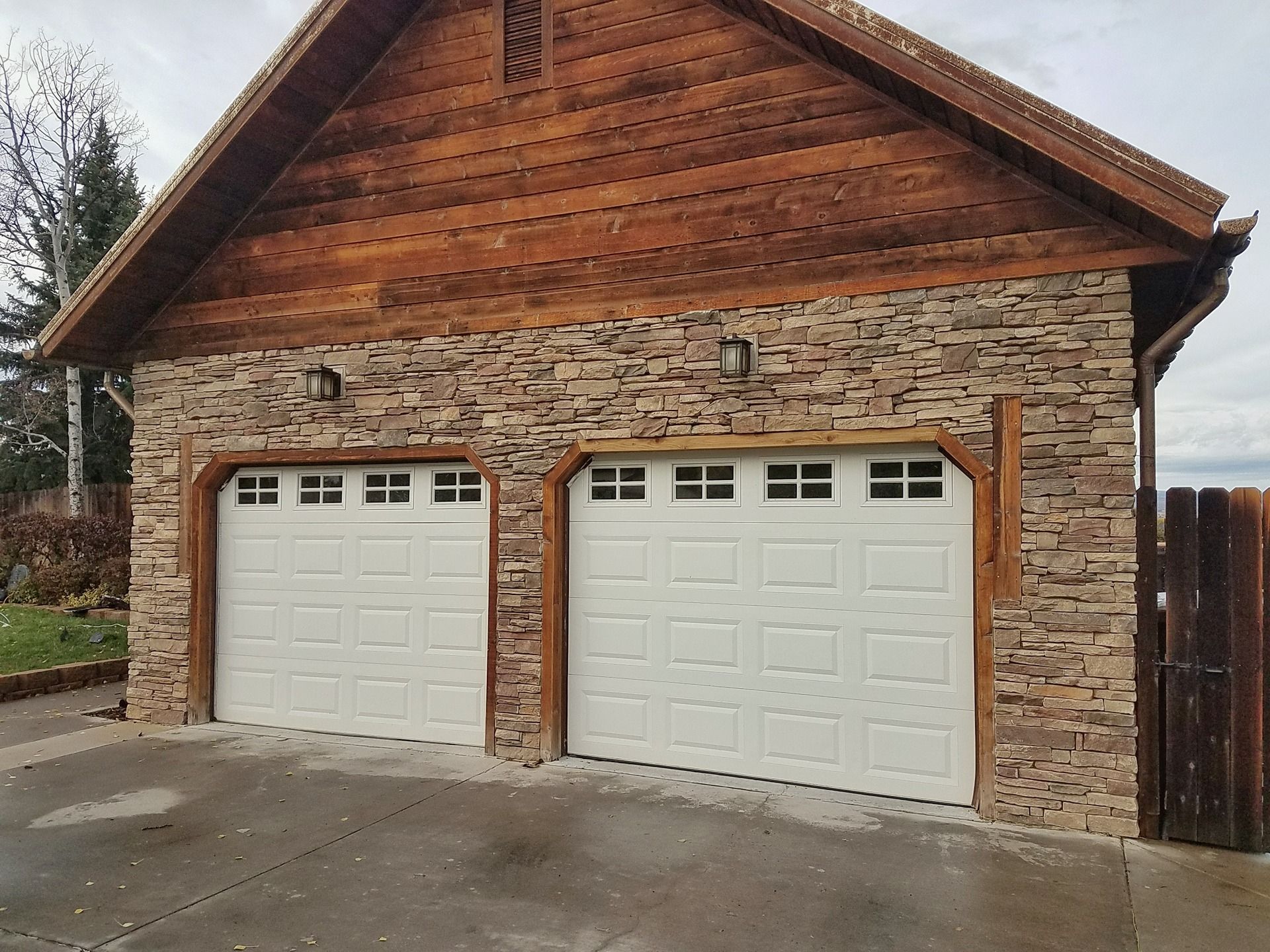 A two-car garage with white paneled doors, stone siding, and a gabled wooden roof set against a cloudy sky.