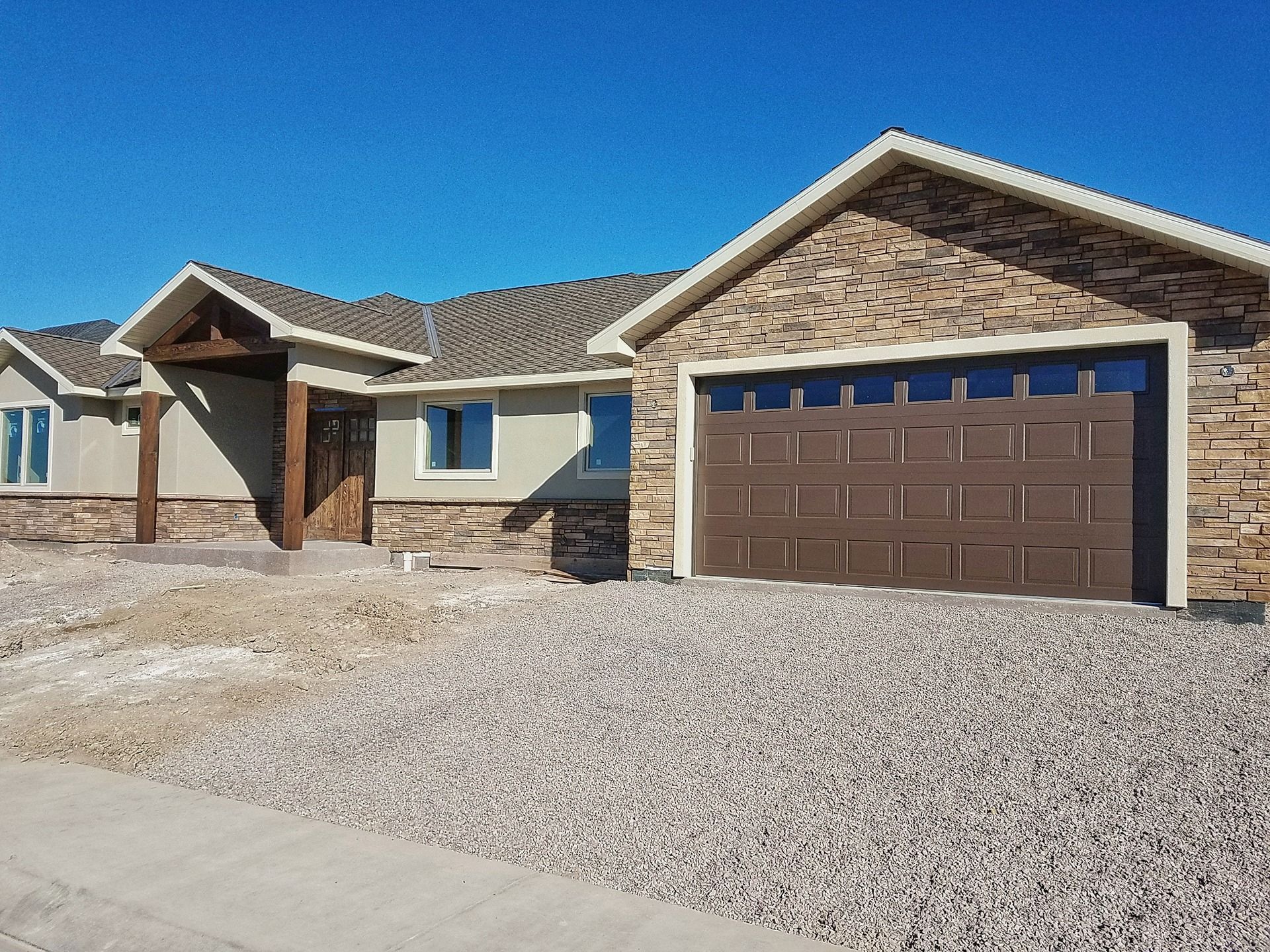 A single-story ranch house with tan stone siding, a dark brown garage door, and a covered front entryway under a blue sky.