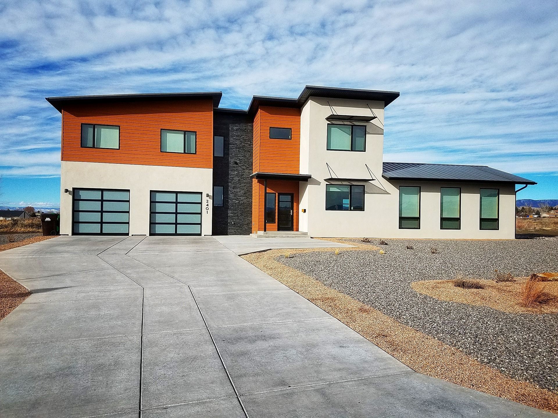 Modern two-story house with wood-paneled and stucco siding, a stone center, dual garage doors, and a gravel yard.