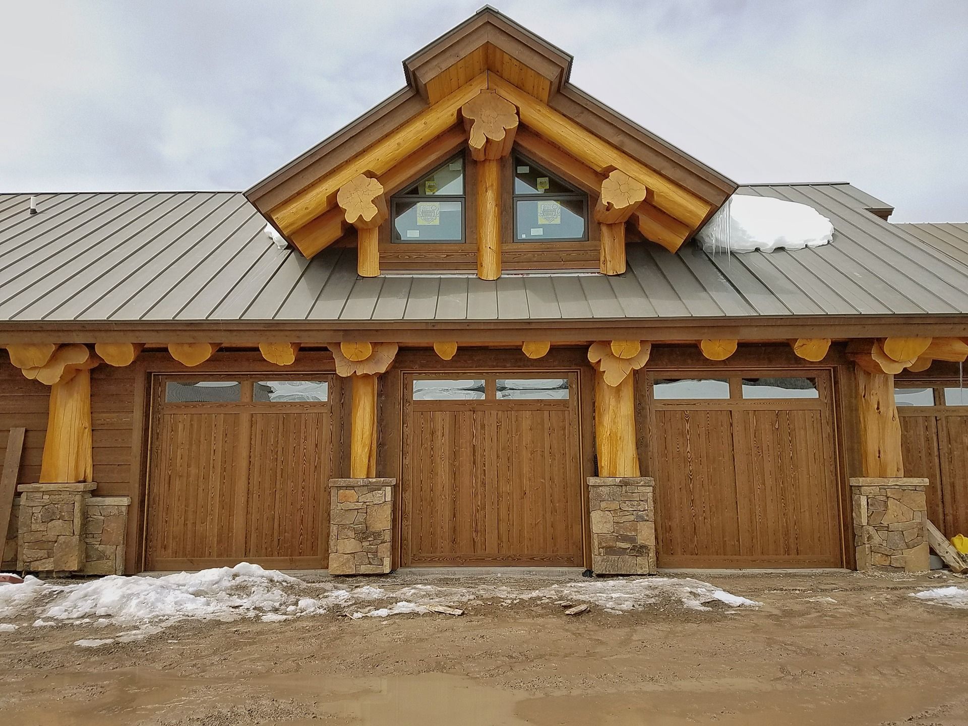 A rustic log cabin-style garage with three wooden doors, stone pillars, and a dormer window under a metal roof in snow.