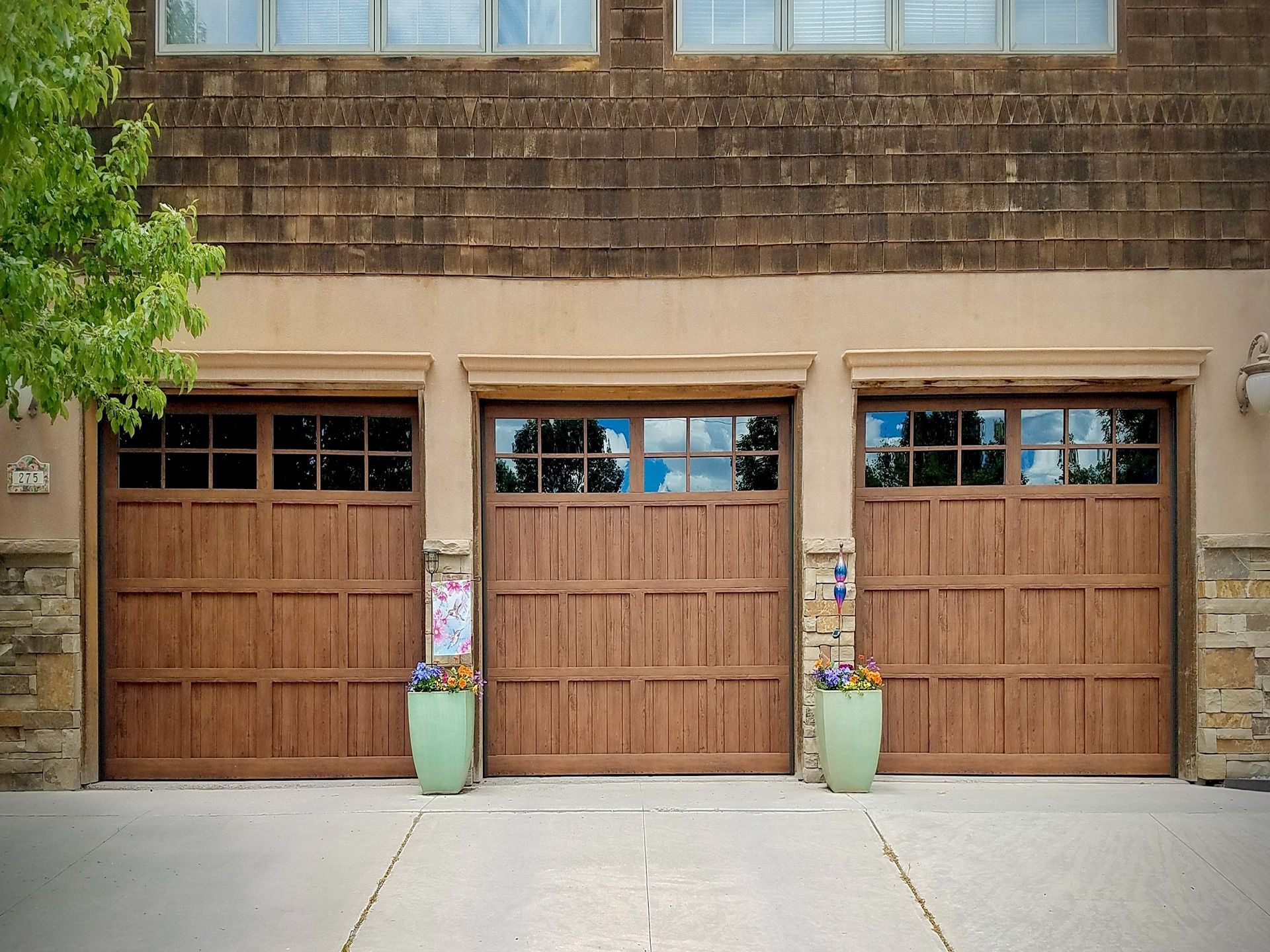 Three wooden garage doors with glass window panels and green planters on the concrete driveway in front of a stucco home.