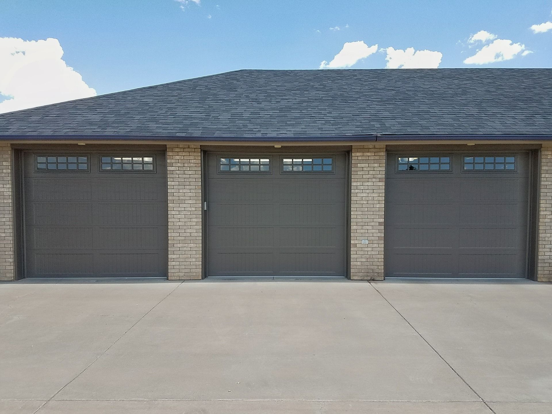 A three-car garage with gray doors featuring windows, set under a dark shingled roof with a concrete driveway in front.
