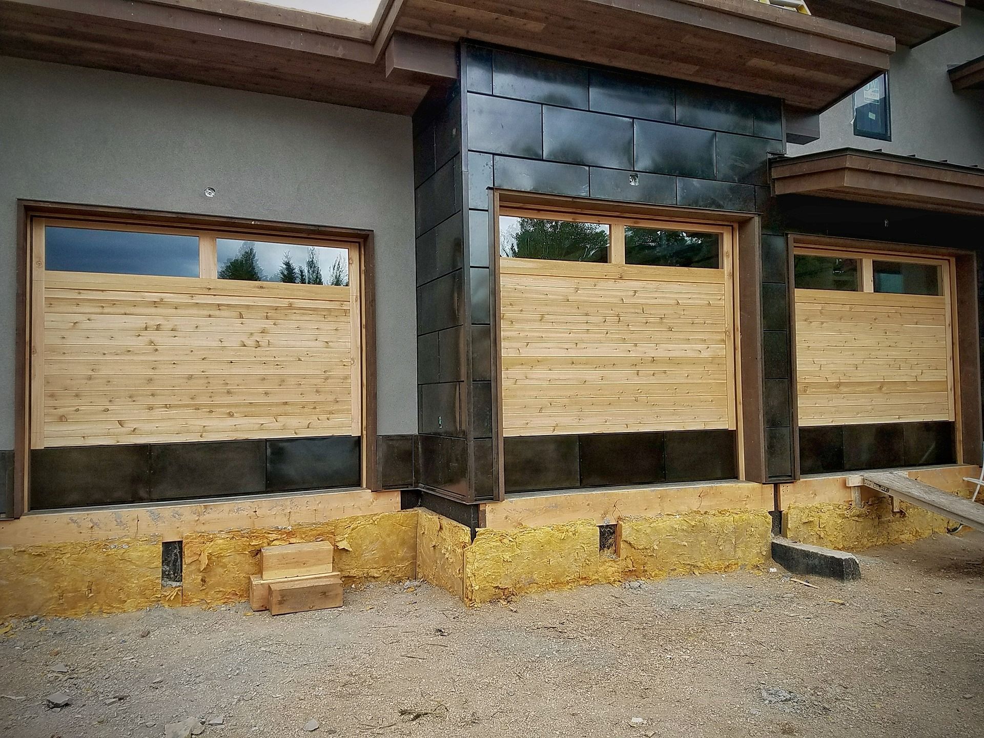 A house exterior under construction, featuring wooden garage doors, black metal siding accents, and exposed foundation.