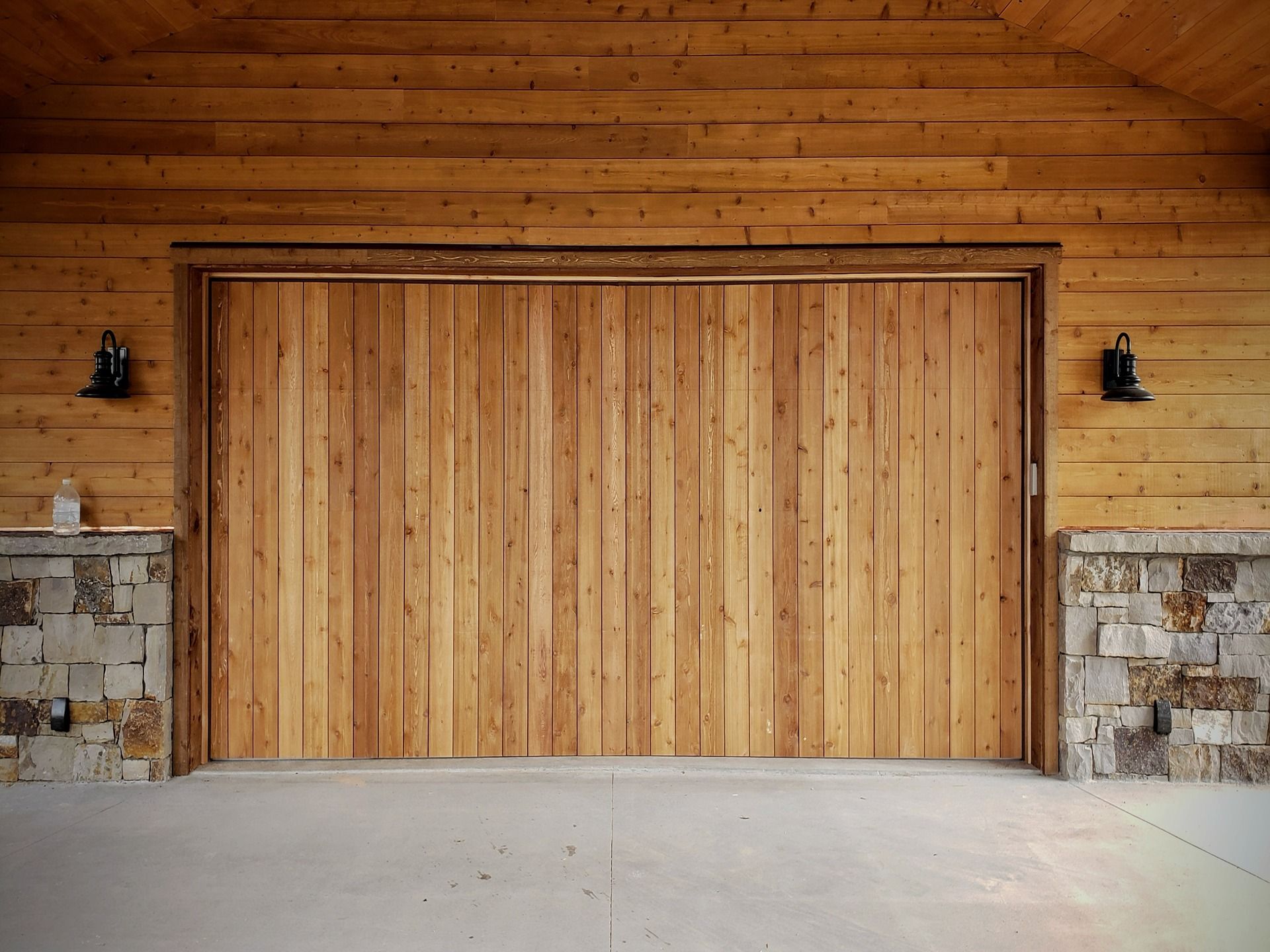 A closed garage door made of vertical wood planks, flanked by stone walls and wood siding, centered in a rustic garage.