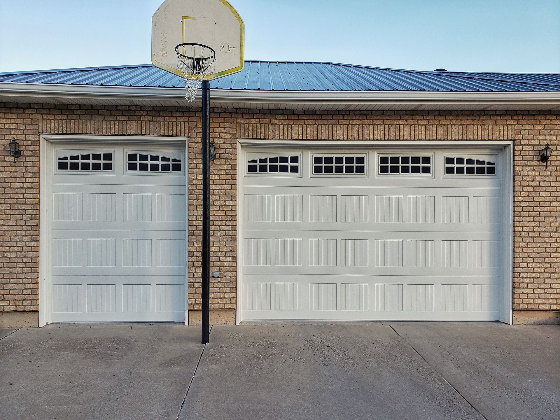 A brick garage with one single and one double white paneled door, beneath a metal roof, with a basketball hoop in front.