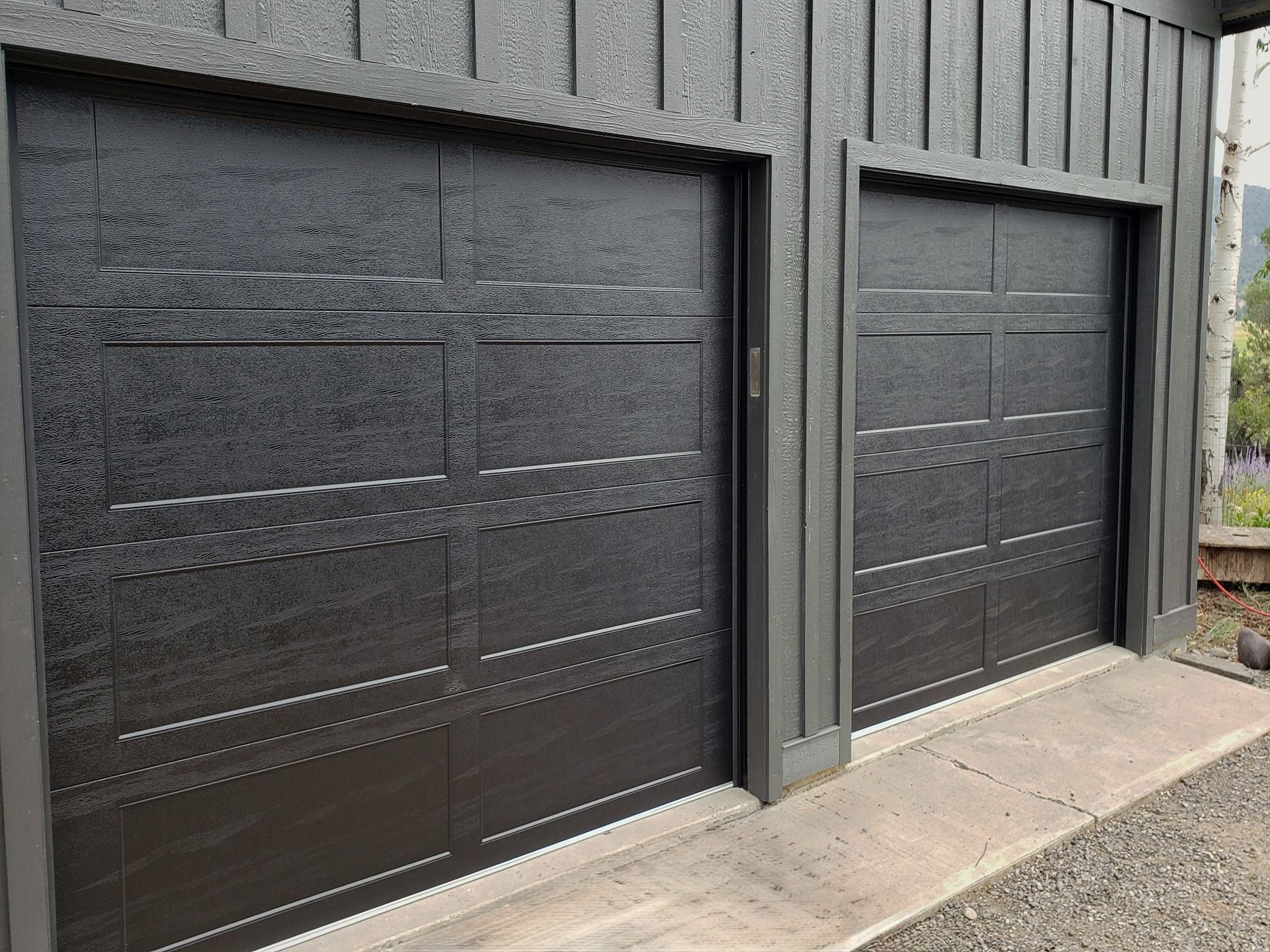 Two dark gray garage doors with a horizontal panel design, installed on a building with vertical wood siding.