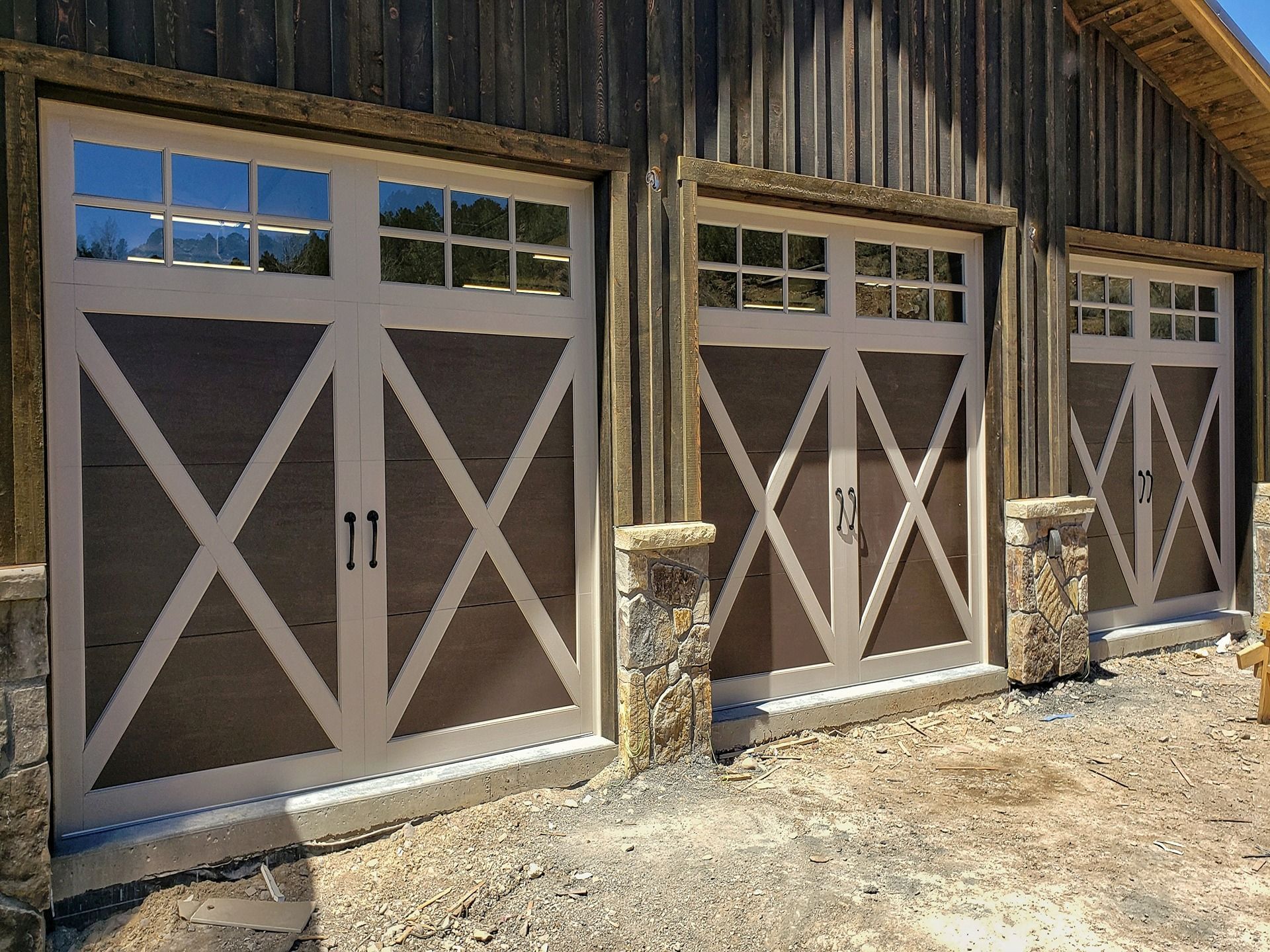 Three rustic, light-colored carriage-style garage doors with X-bracing and windows set against a dark wood building.
