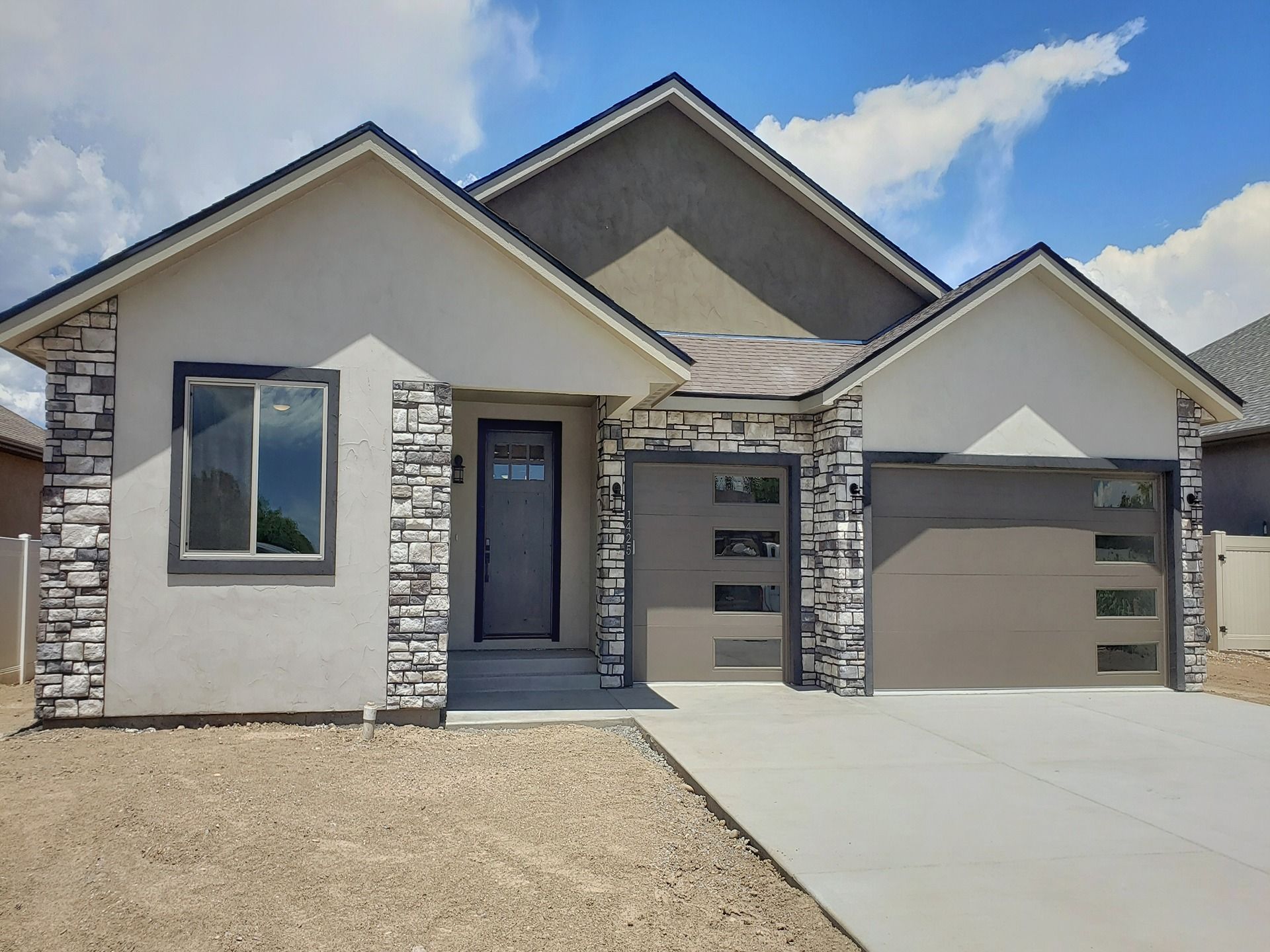 A single-story suburban house with beige stucco, gray stone accents, a dark front door, and a two-car garage.
