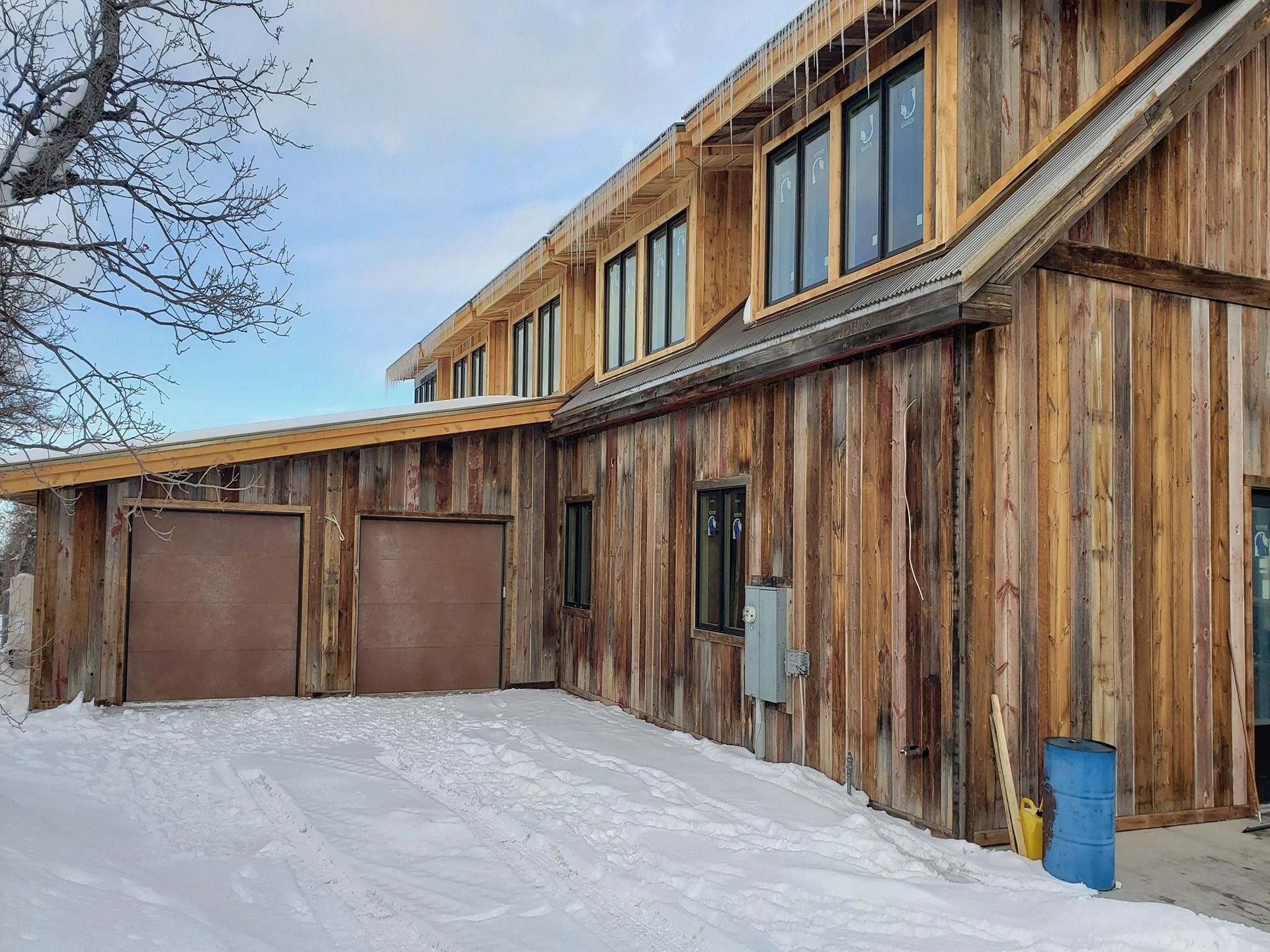 A rustic wooden building with two garage doors and upper-level windows sits on a snowy ground under a blue winter sky.