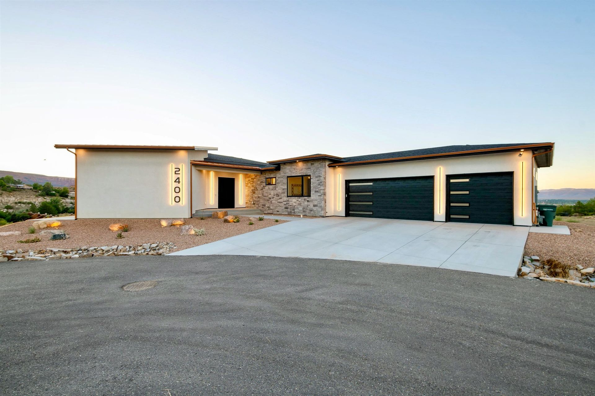 A single-story modern home with white stucco, stone accents, two dark garage doors, and a concrete driveway at dusk.