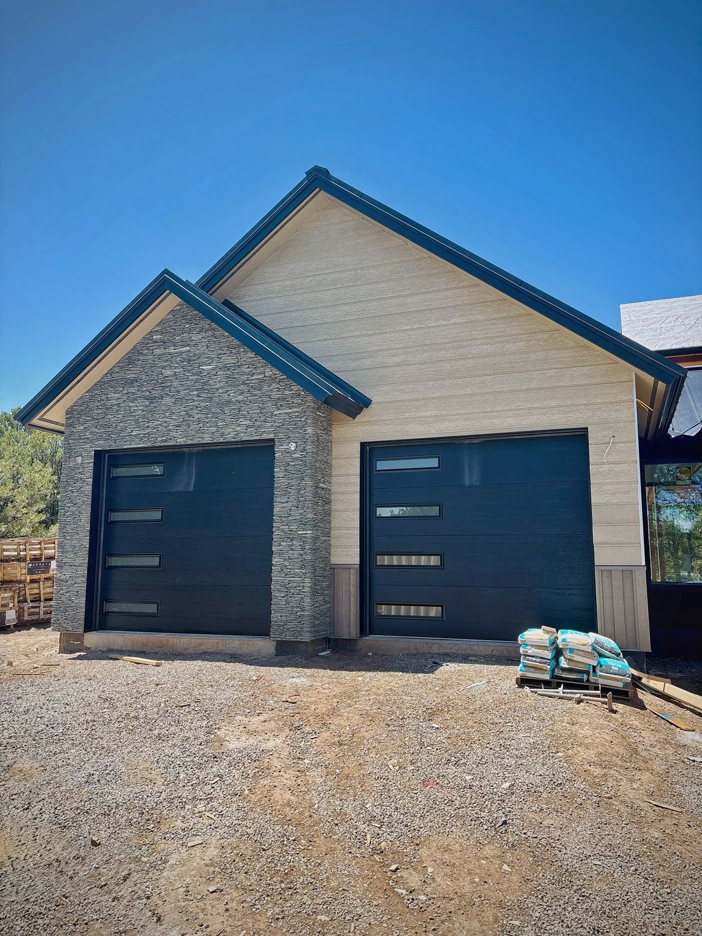A new house garage under construction with stone siding, cream walls, and two modern black doors under a blue sky.
