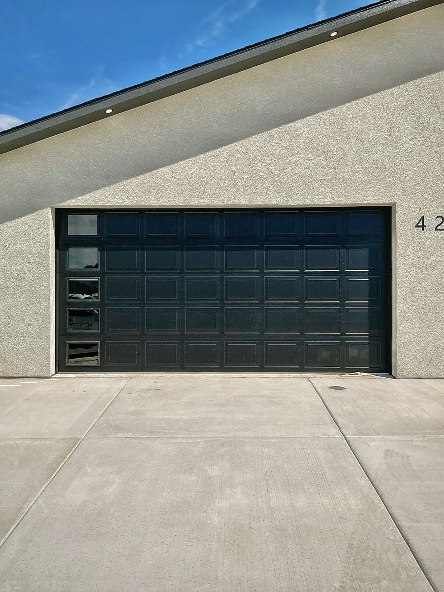 A black garage door with a vertical row of five windows on the left, installed in a light-colored stucco exterior wall.