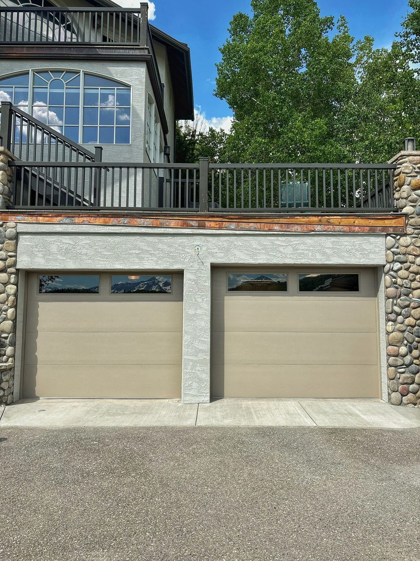 Two tan garage doors under a deck with stone-textured pillars and a house exterior in a sunny, tree-lined setting.