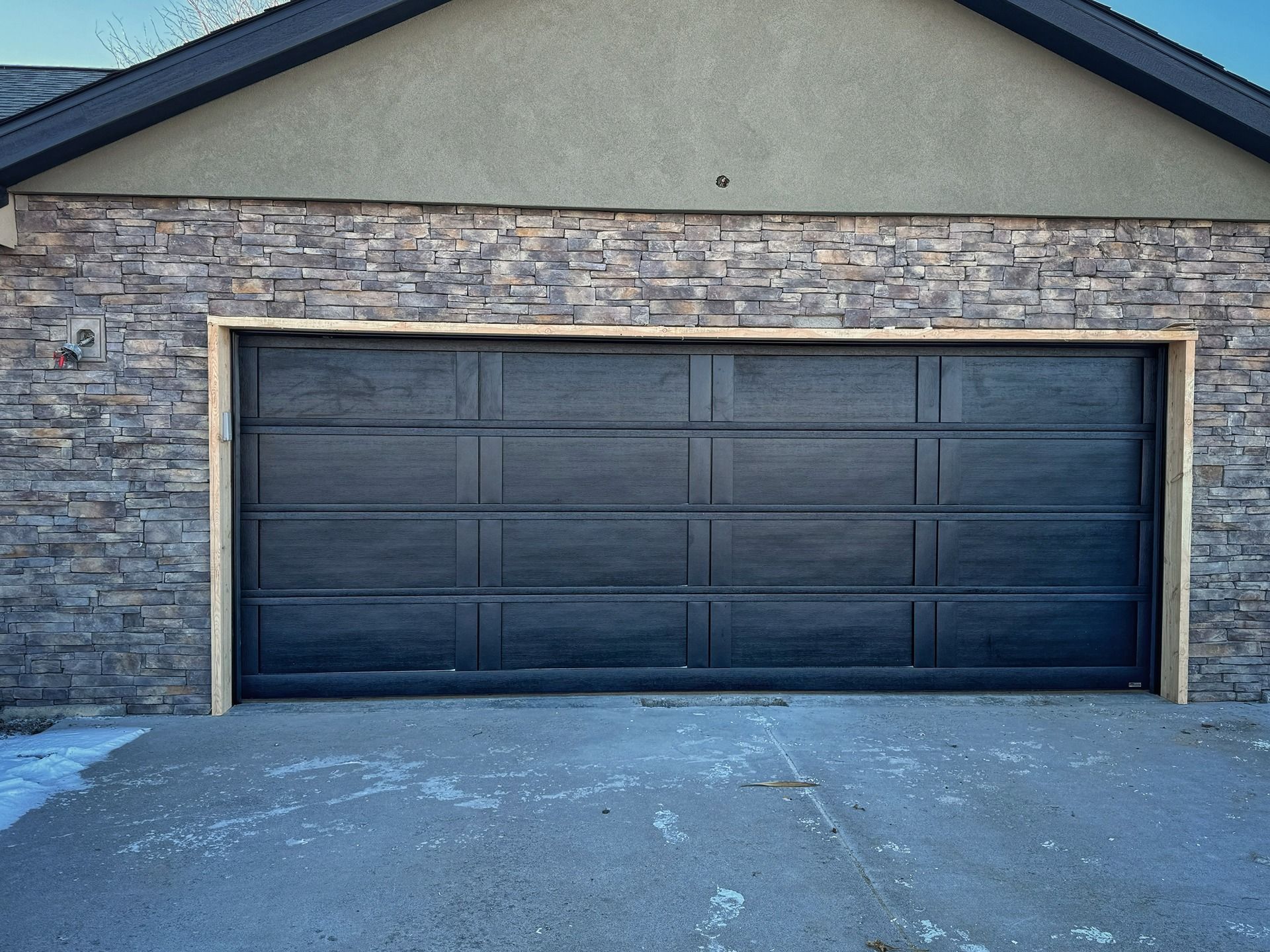 A dark, multi-panel garage door framed in light wood trim, set against a stone-veneer wall.