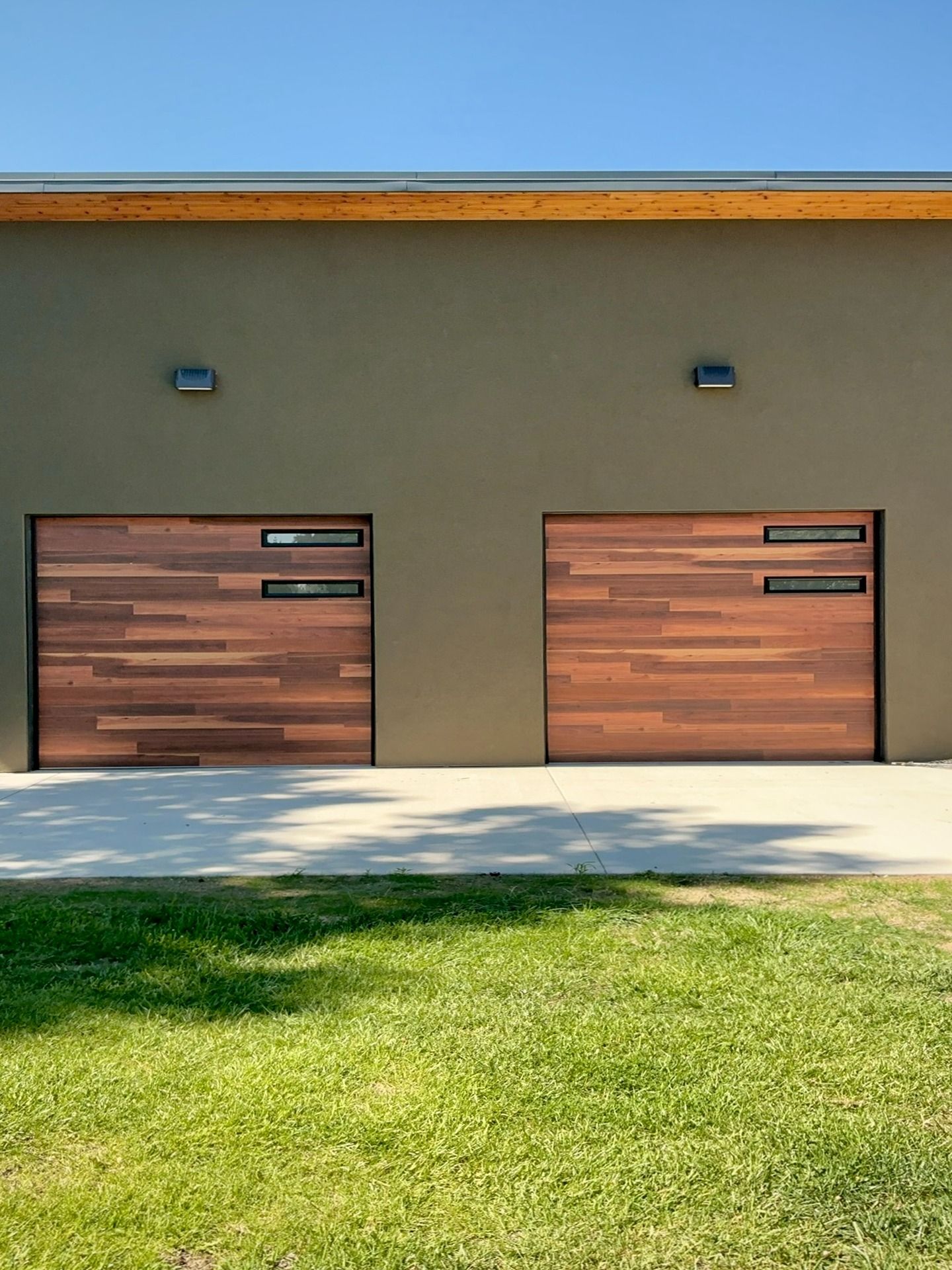 Two wooden garage doors with horizontal slats and thin, horizontal windows, set into a plain olive wall under blue sky.