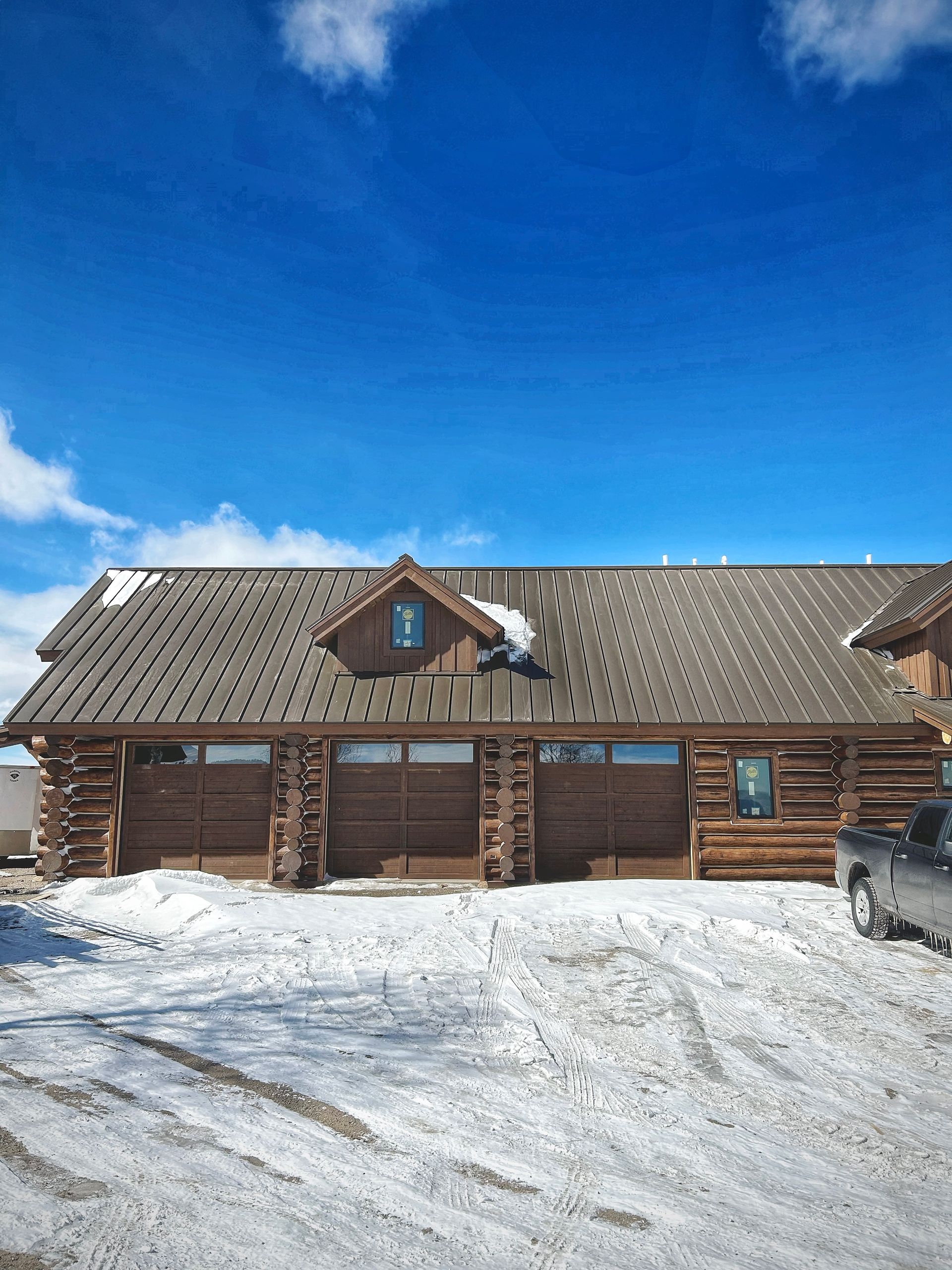 Snow-covered log cabin with a gray roof under a bright blue sky, with a vehicle parked on the right.