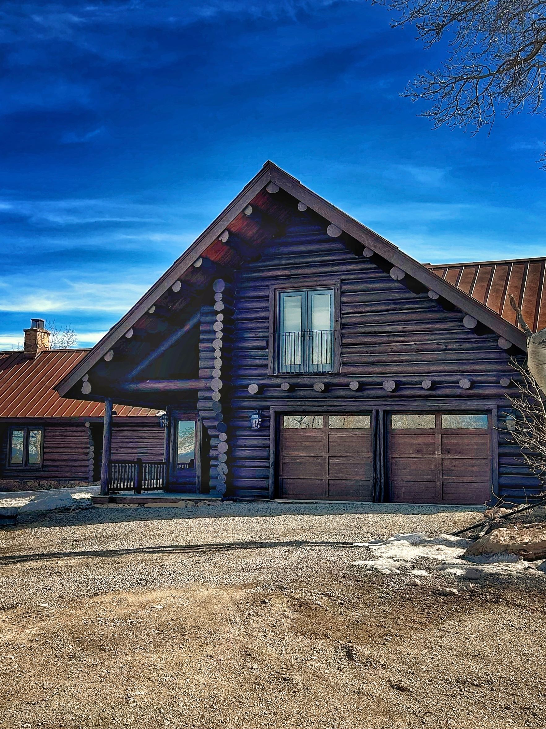 Rustic log cabin with garage doors under a bright blue sky on a snowy, gravel lot
