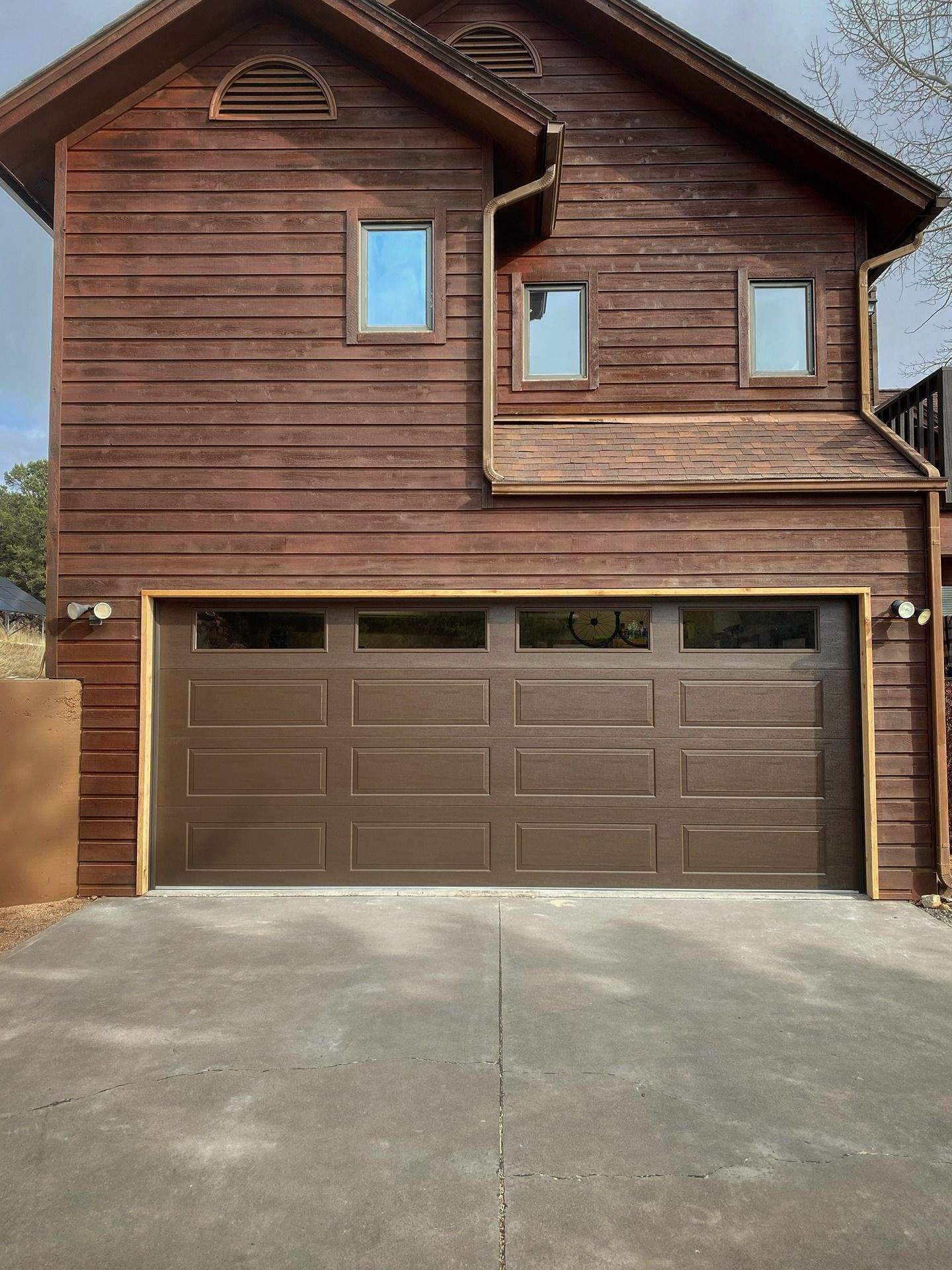 A two-story house with dark brown wood siding and a prominent single-car garage door, viewed from a concrete driveway.