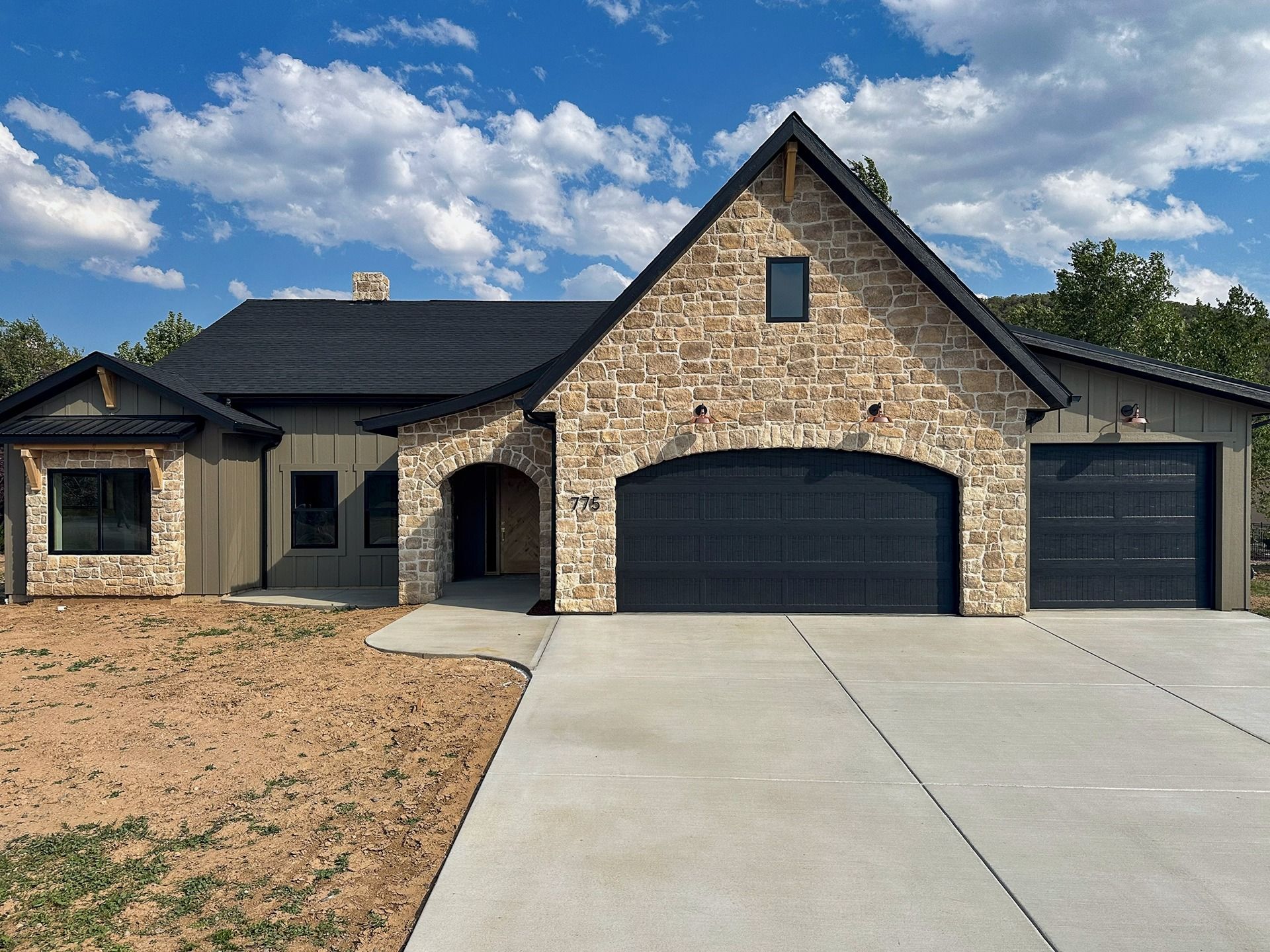 A single-story stone house with a dark roof and two garage doors, set behind a concrete driveway under a blue sky.